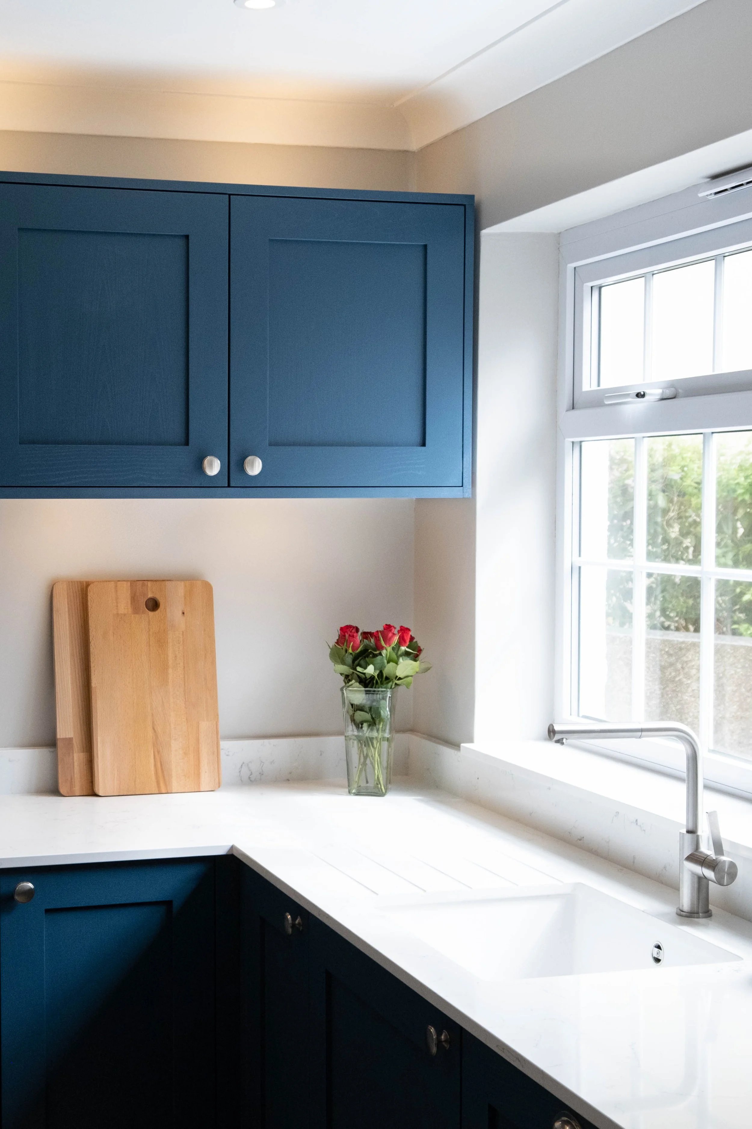 A kitchen with blue upper cabinets, a white countertop with a built-in sink, a window letting in natural light, a glass vase with red roses, and two wooden cutting boards leaning against the wall.