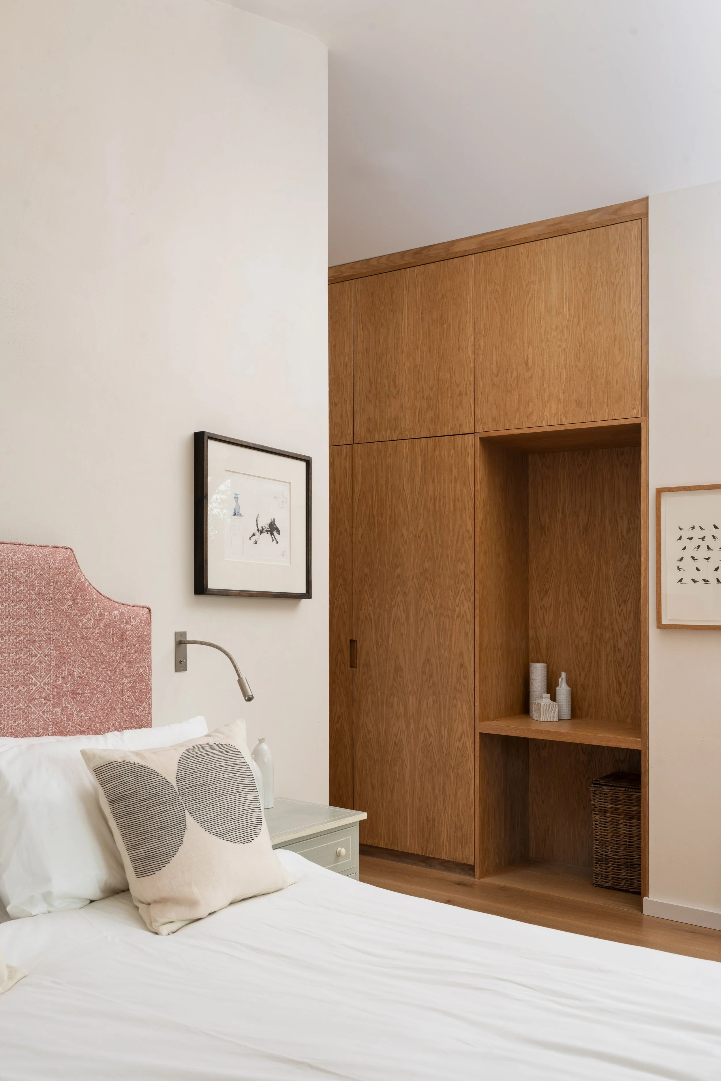 A bedroom with white walls, a bed with white sheets and pillows featuring black geometric patterns, a nightstand with a lamp, and a wooden built-in bookshelf with decorative objects.