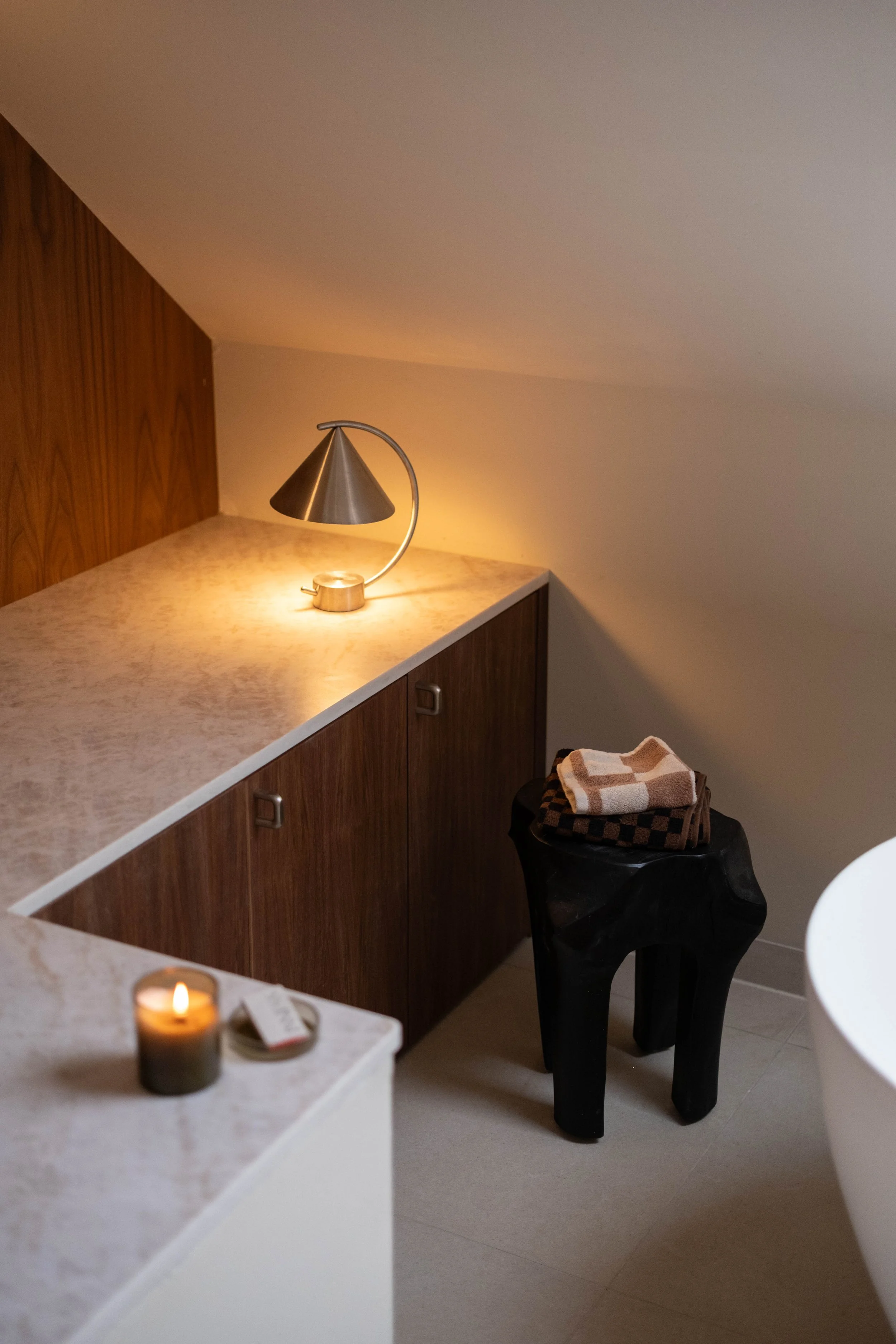 A lit candle on a white surface in the foreground, a countertop with a small lamp and an orange candle in the background, a black decorative stool with folded towels on top, and a section of a white chair or table to the right.