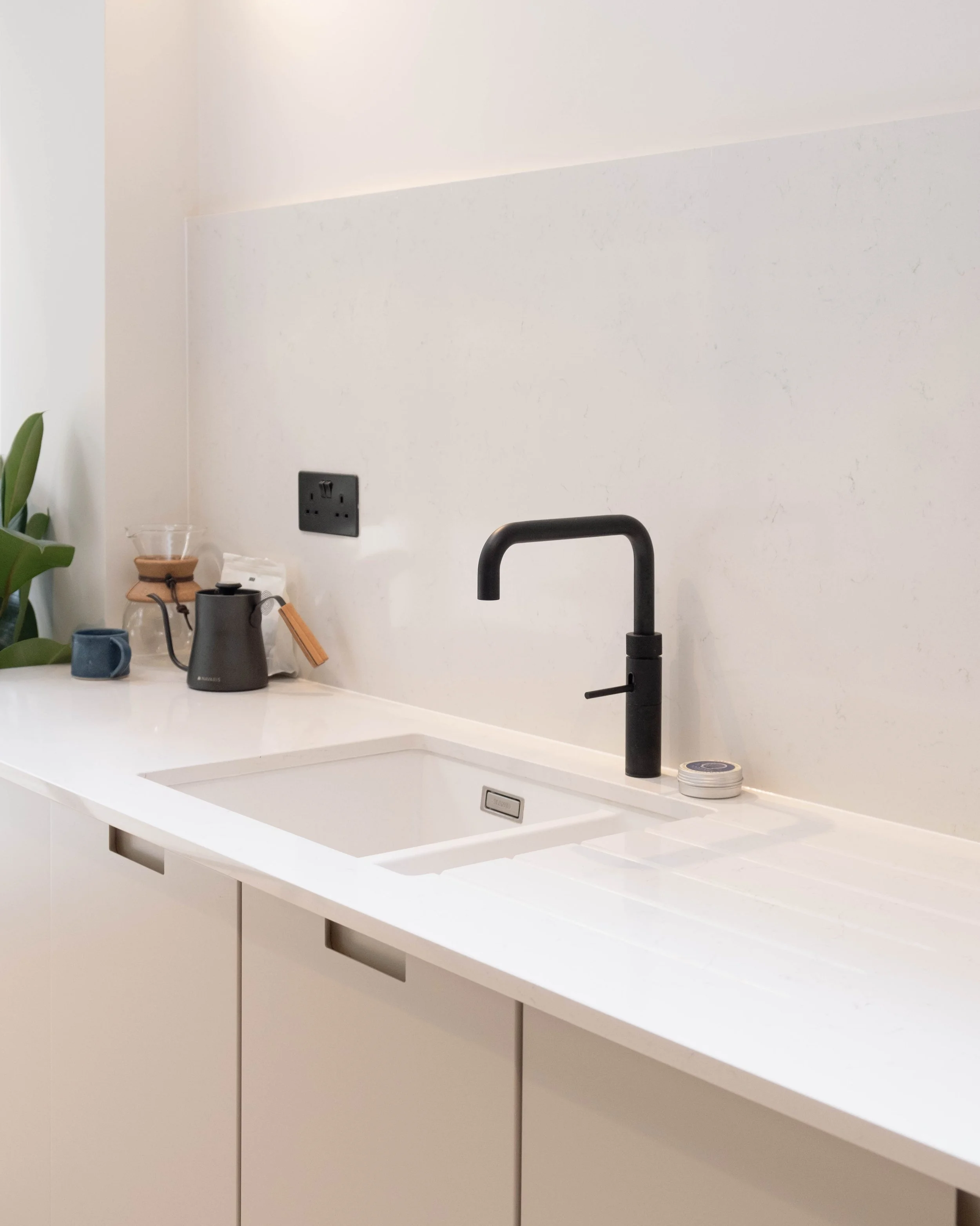 Modern white kitchen countertop with black faucet, black electrical outlet, a potted plant, coffee maker, cups, and a small container on the counter.