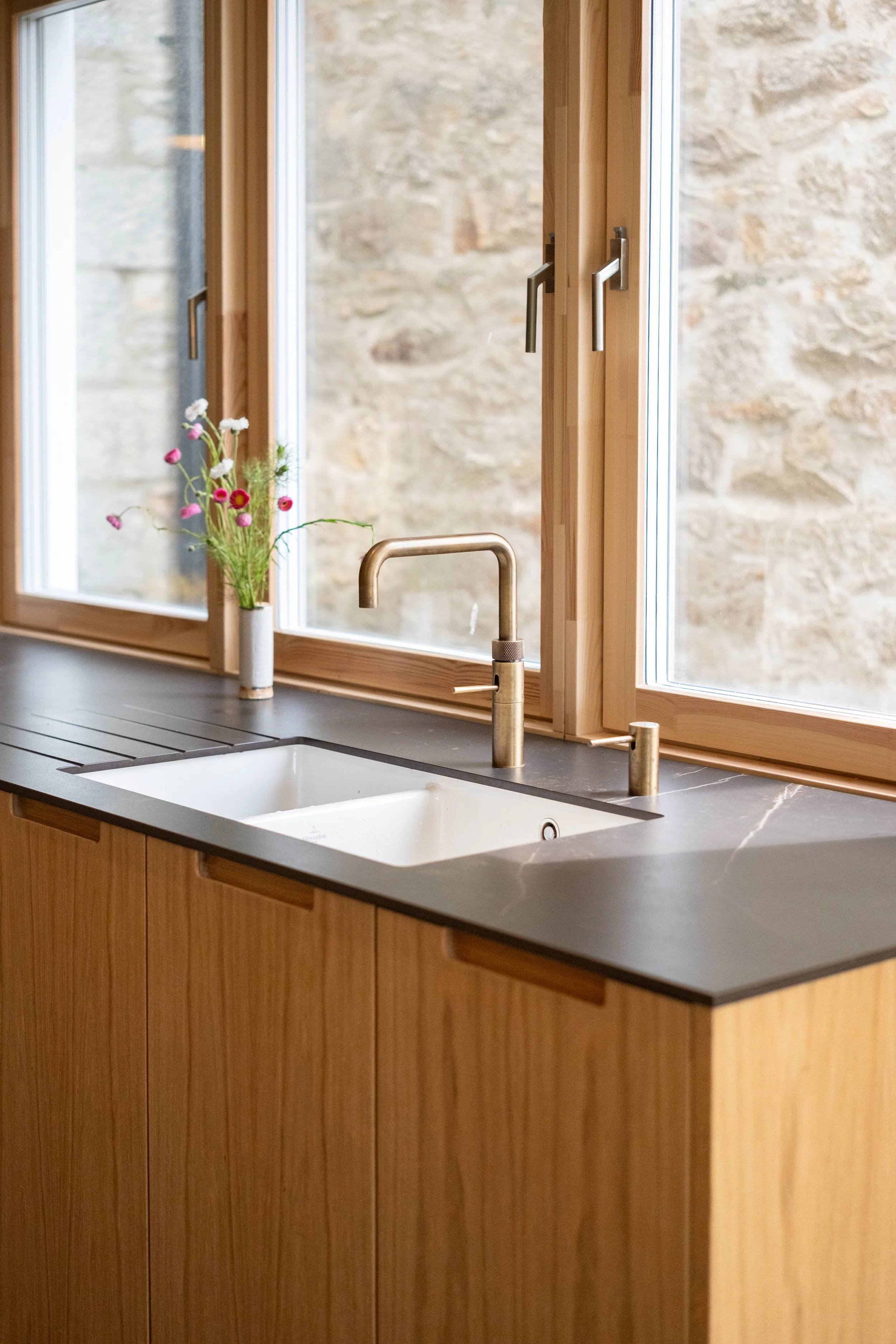 A kitchen countertop with a white sink, a garden-style faucet, and a small vase with pink and white flowers on a window sill. The window has wooden frames and offers a view of a stone wall outside.