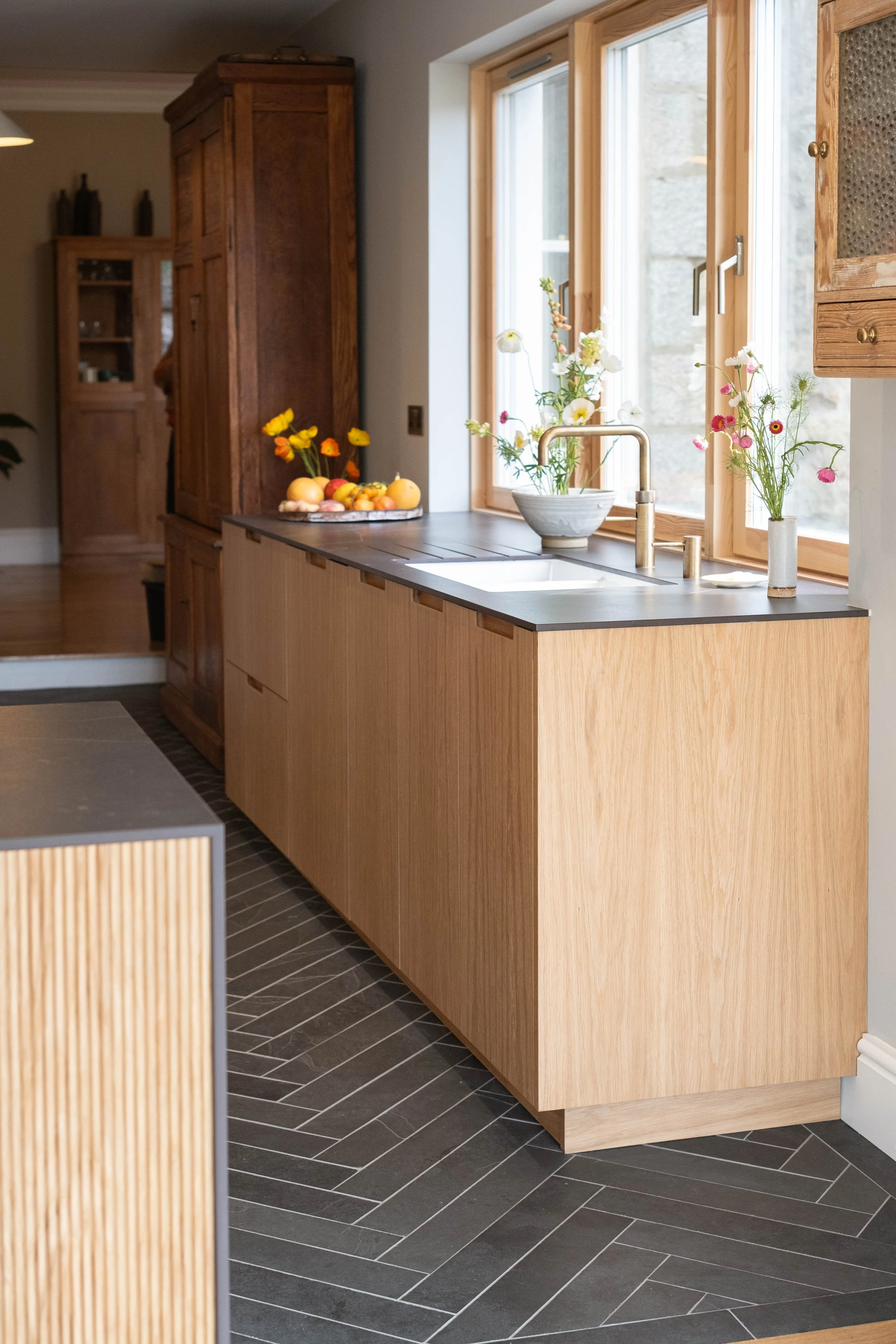 A kitchen with light wooden cabinets and a countertop with a bowl of flowers, a stony window with a faucet, and a dark tiled floor.