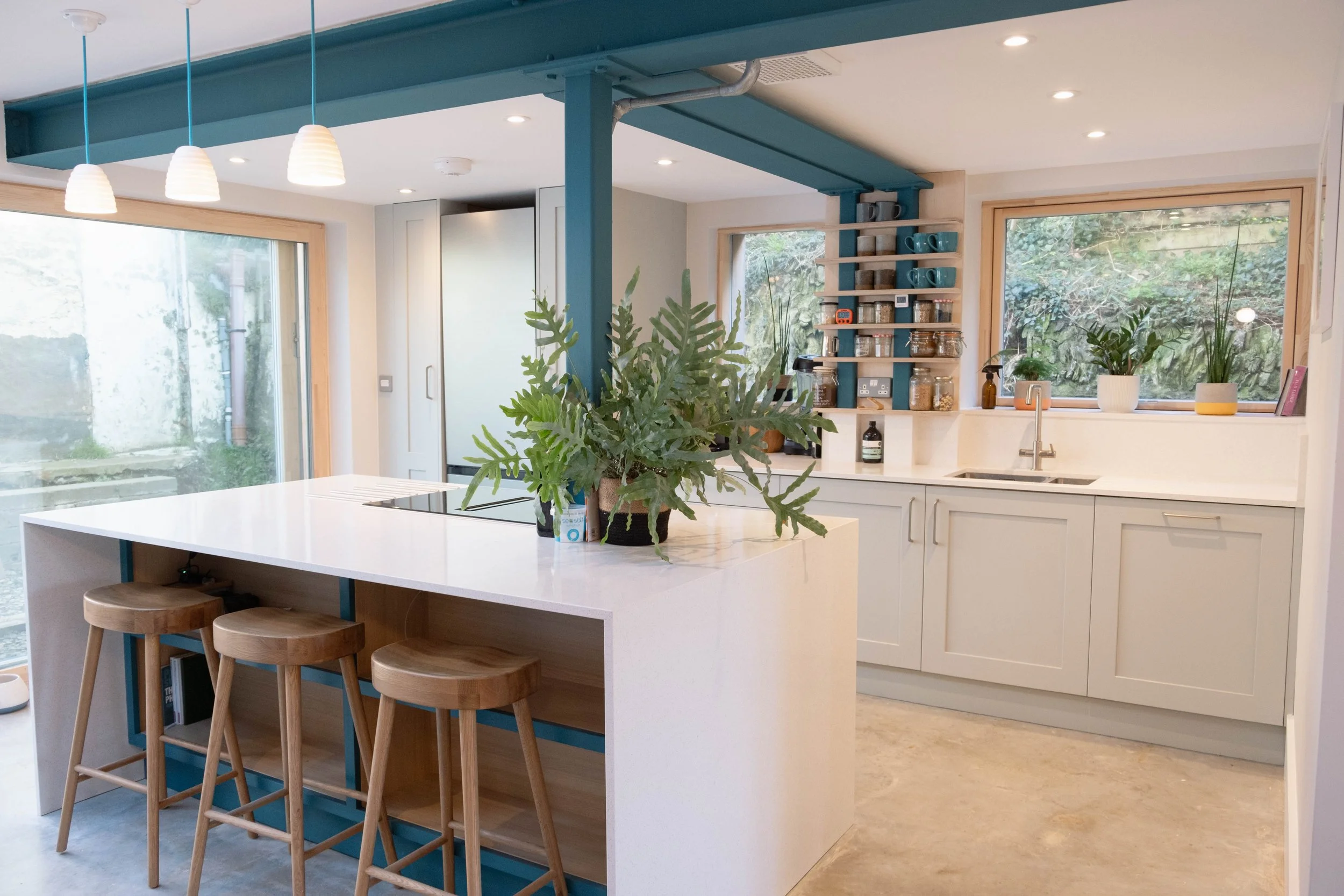 Modern kitchen with white island counter, wooden bar stools, large window with greenery outside, open shelving with dishes and jars, plants on the counter, and pendant lights hanging from the ceiling.