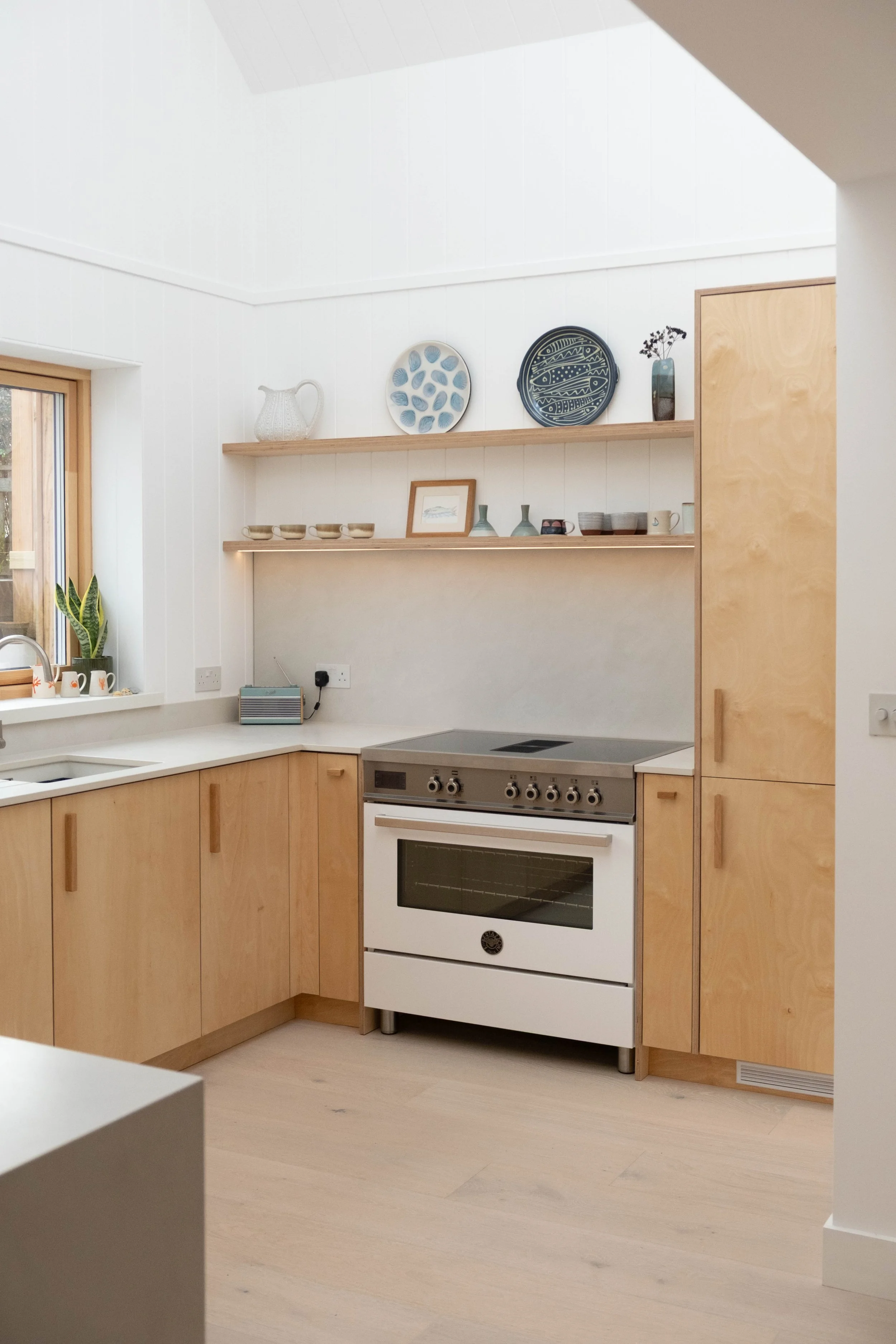 A modern kitchen with white walls, wooden cabinets, a stainless steel oven, open wooden shelves with decorative plates, bowls, and vases, a window with a plant, and light-colored flooring.