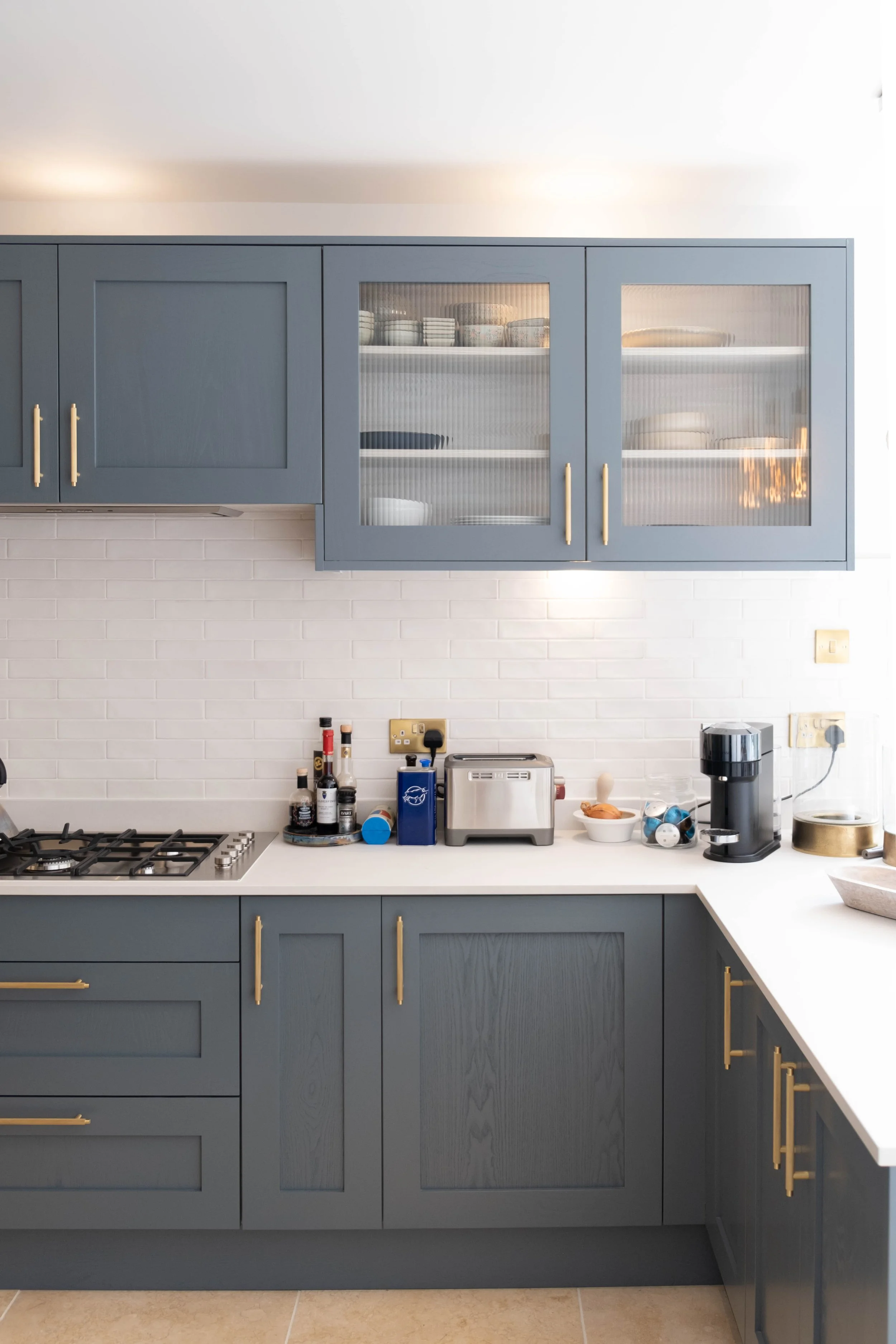 A modern kitchen with blue cabinets, some with glass doors, white countertop, and various small appliances and bottles on the counter. There's a gas stove, toaster, coffee machine, and a bowl.