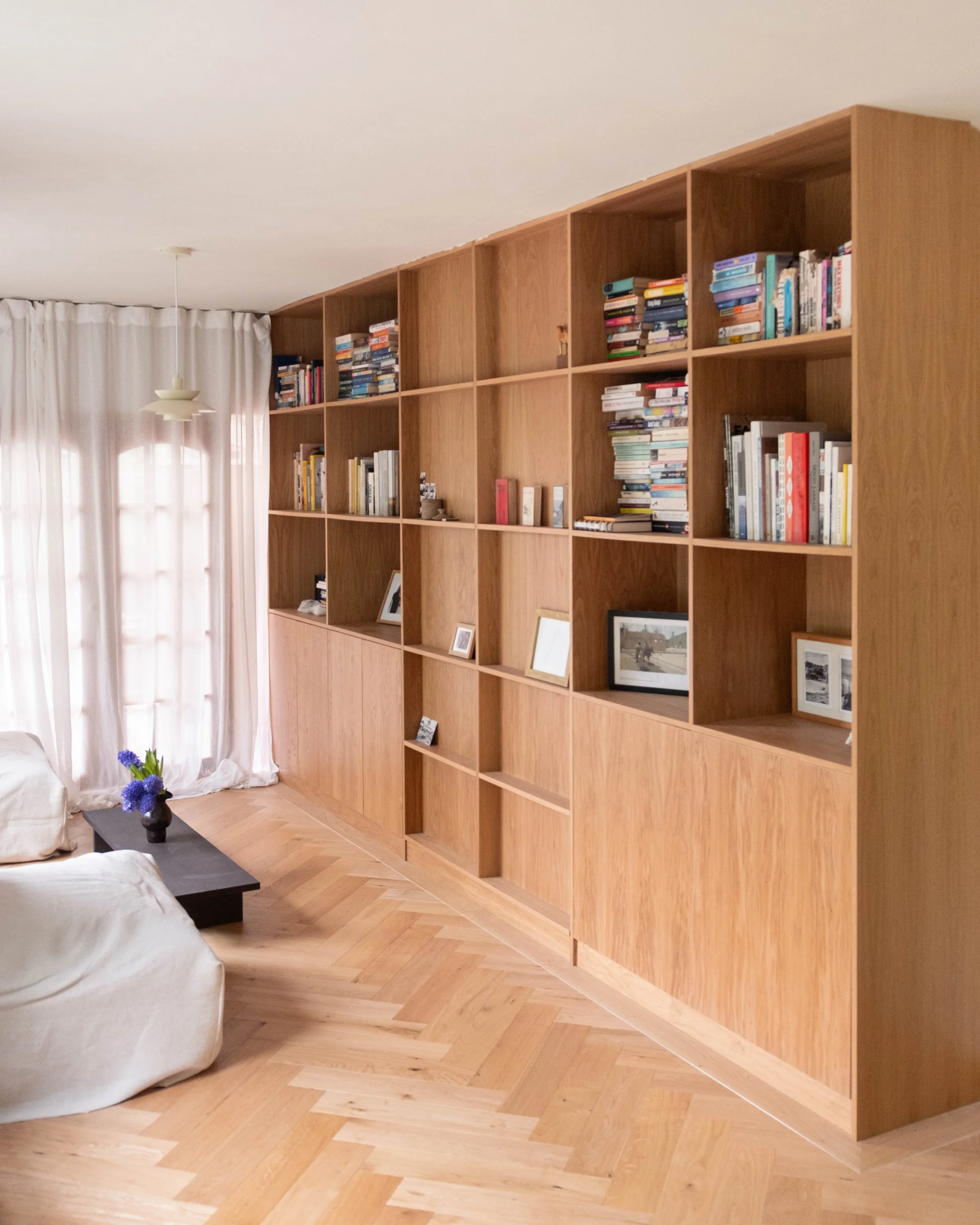 Living room with a large wooden bookshelf filled with books and framed photographs, a white curtain covering large windows, a white sofa, a black coffee table with a vase of purple flowers, and a hanging ceiling light.