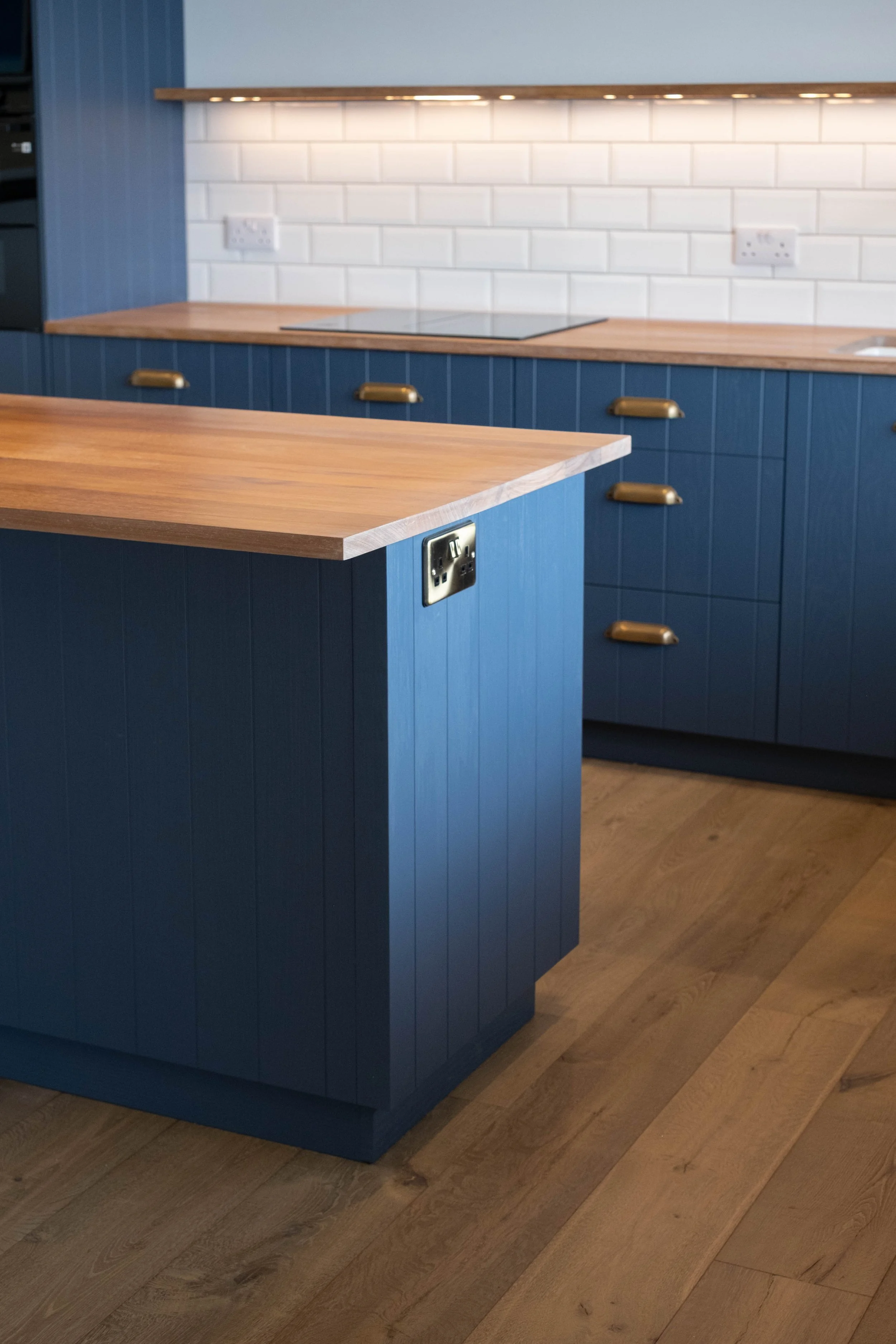 A kitchen with blue cabinetry and a wooden countertop. There are electrical outlets on the white tiled backsplash and a wooden shelf with lighting above the counter. The floor is wooden.