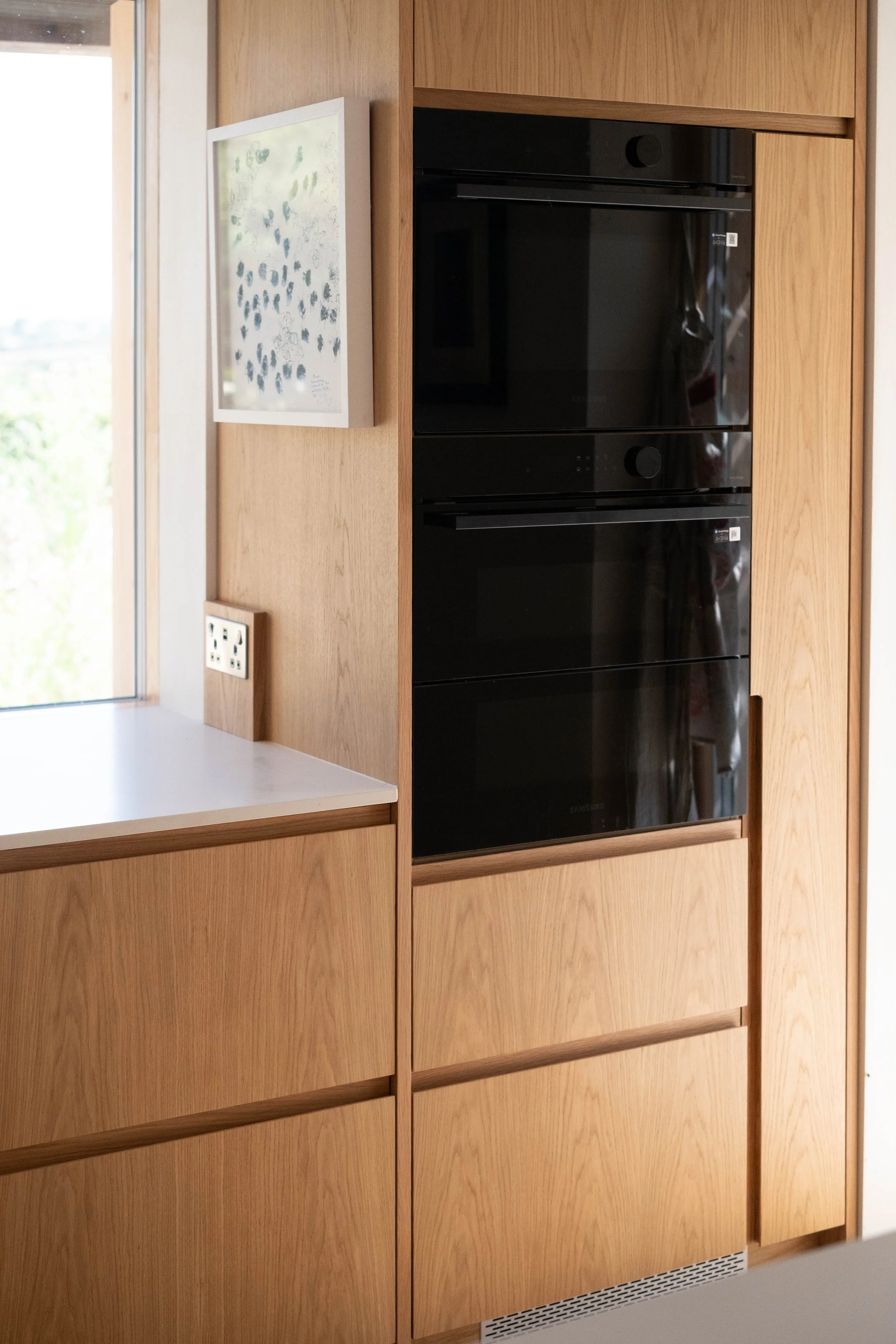 Built-in wooden kitchen cabinets with a black oven and a white countertop. There is a piece of artwork on the wall and a power outlet near the counter.