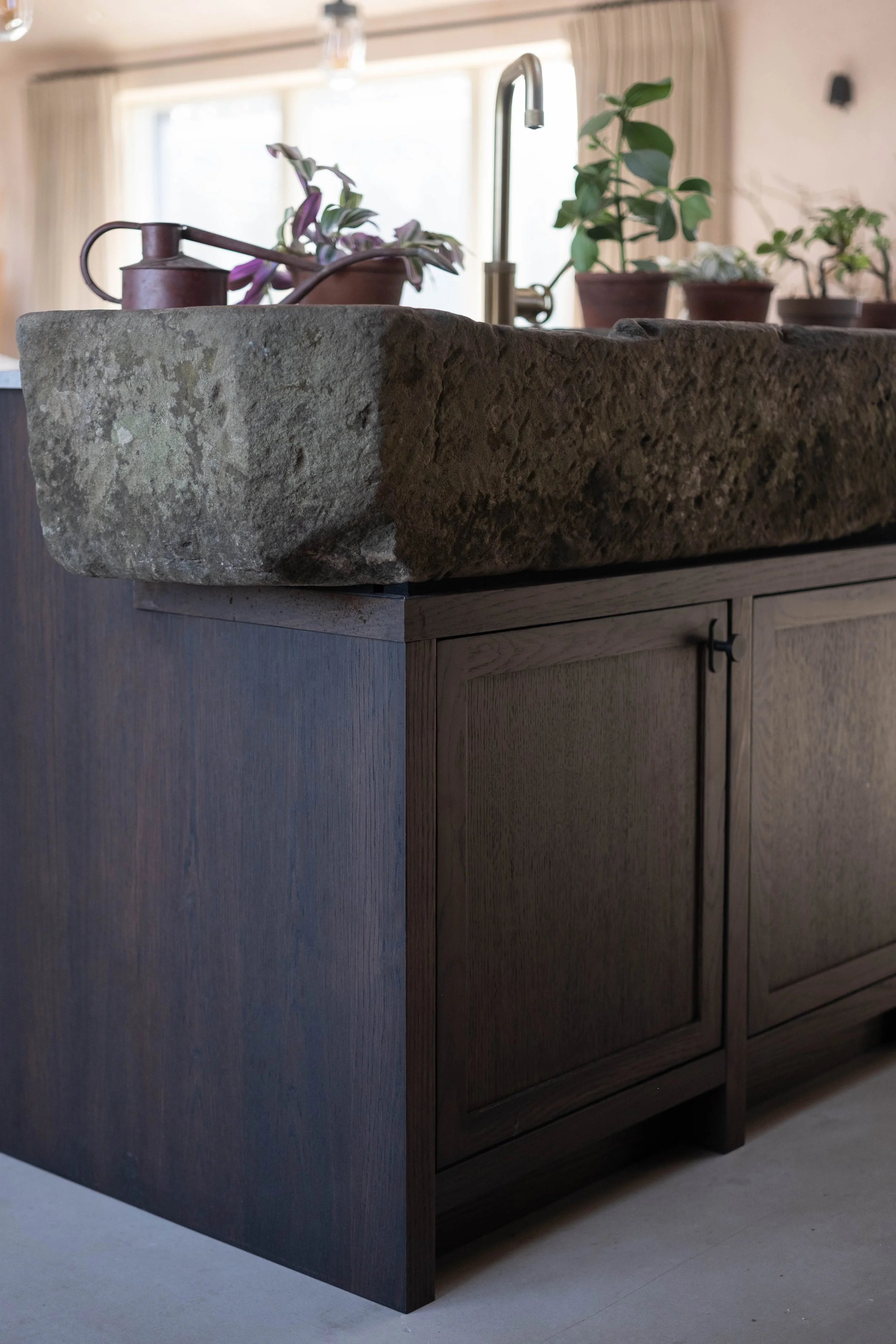 Close-up of a rustic stone kitchen sink on a wooden cabinet with a modern silver faucet, potted plants on the countertop, and a window with curtains in the background.