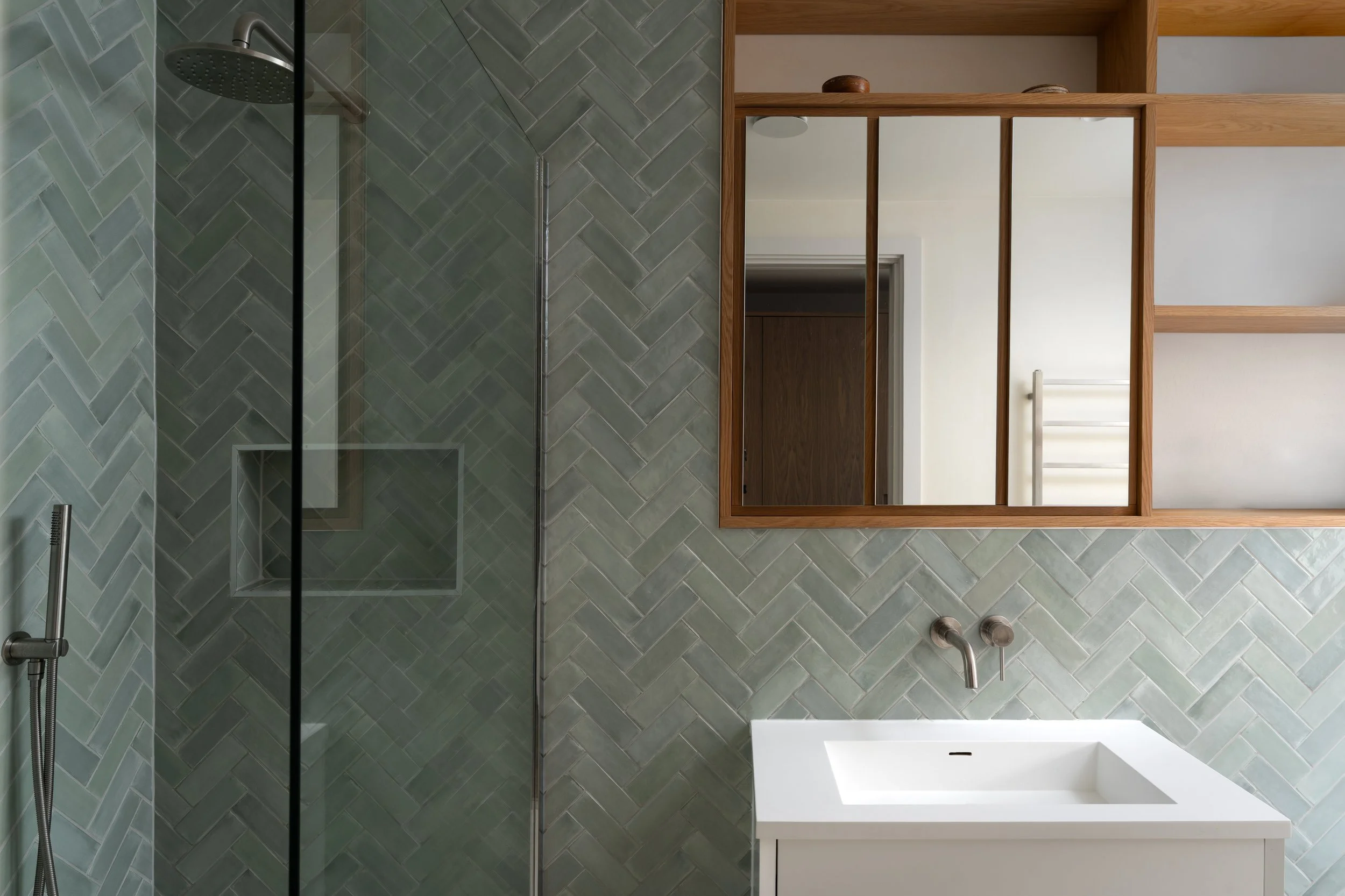 A modern bathroom with a walk-in shower enclosed by glass, a wooden-framed mirror, a white sink vanity, and a white towel rack. The shower features herringbone-patterned green tiles and a rain showerhead. The room has minimalistic decor and a clean, 