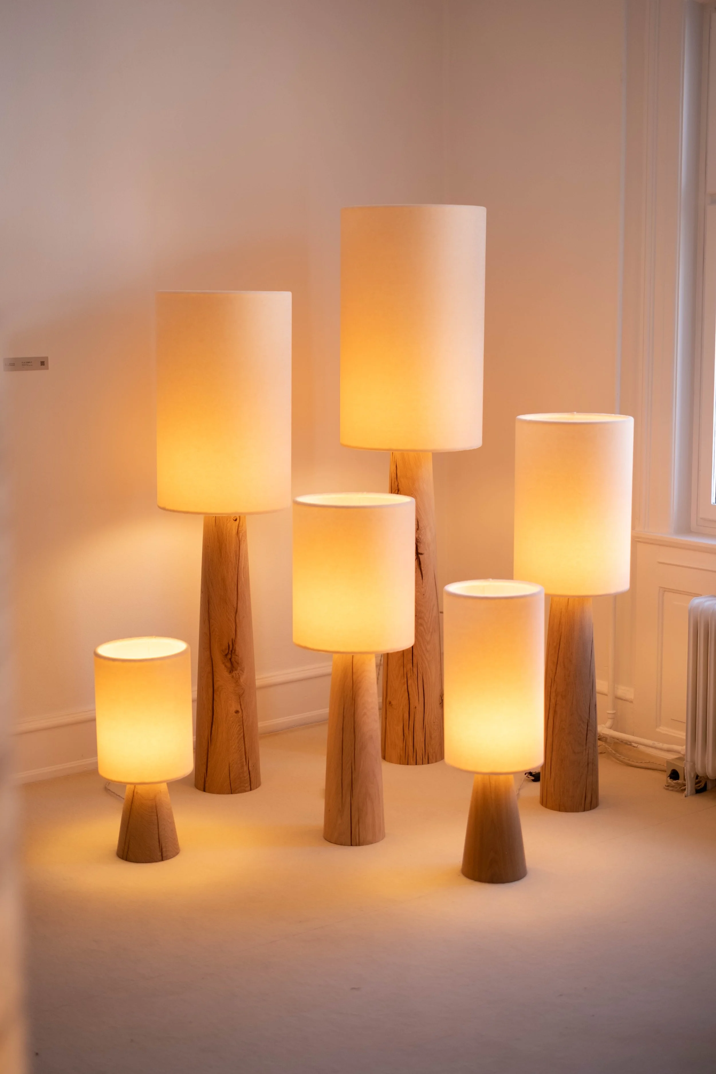 Multiple wooden lamps with cylindrical shades of different heights in a room with beige walls and carpet.
