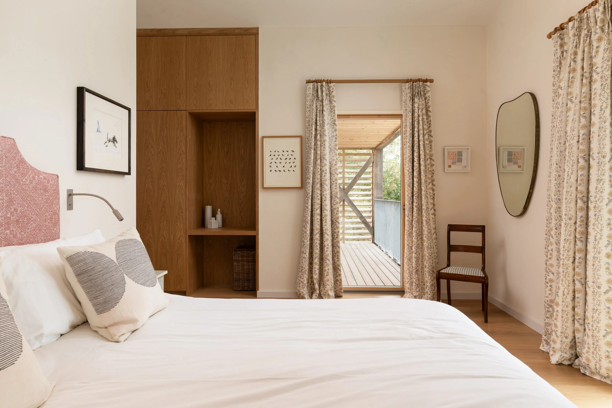 A bedroom with a white bed, pillows, artwork on the walls, wooden cabinets, and a door opening to a balcony with a wooden terrace and greenery outside.