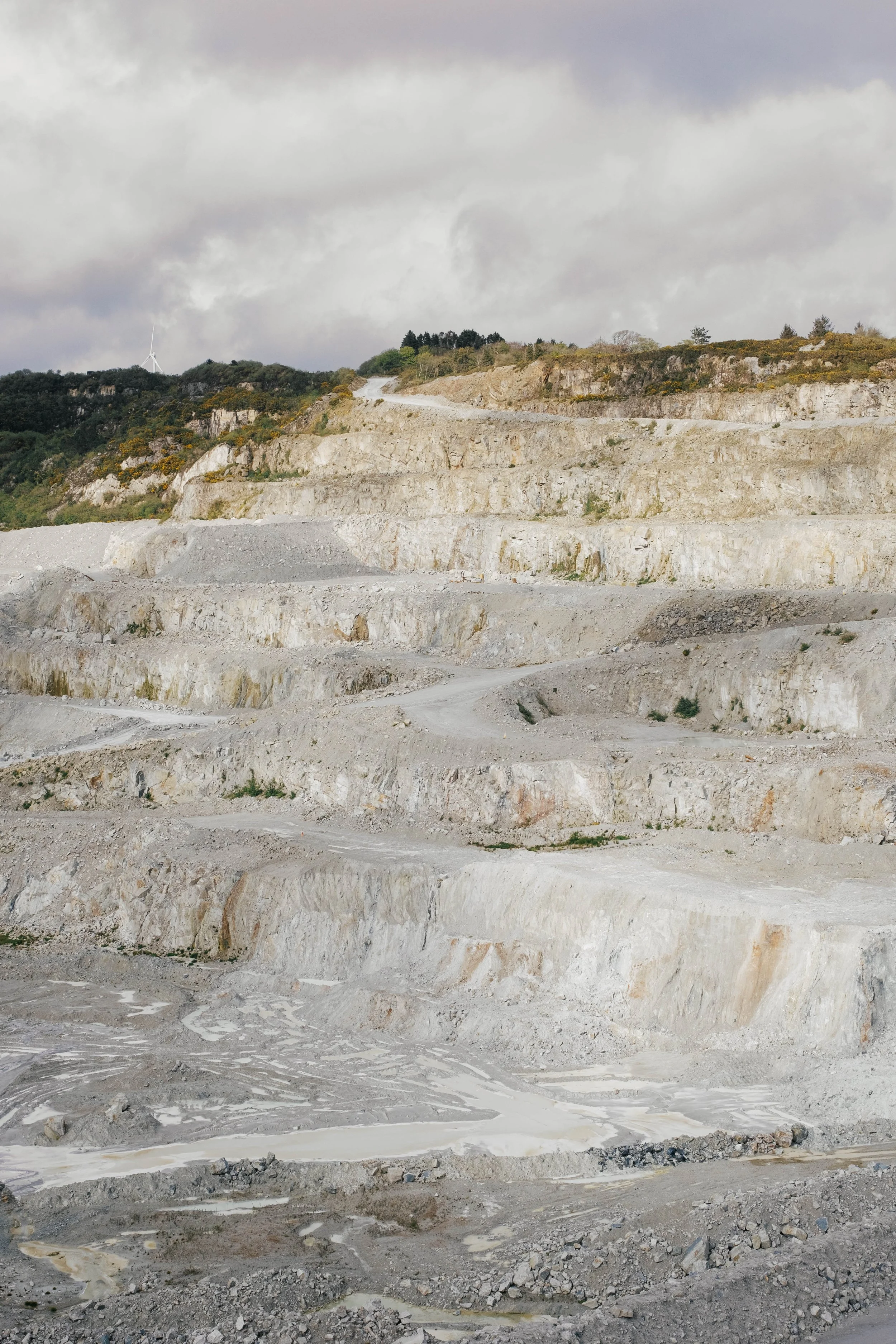 A quarry with multiple terraced steps showing exposed rock and gravel, overcast sky in the background.