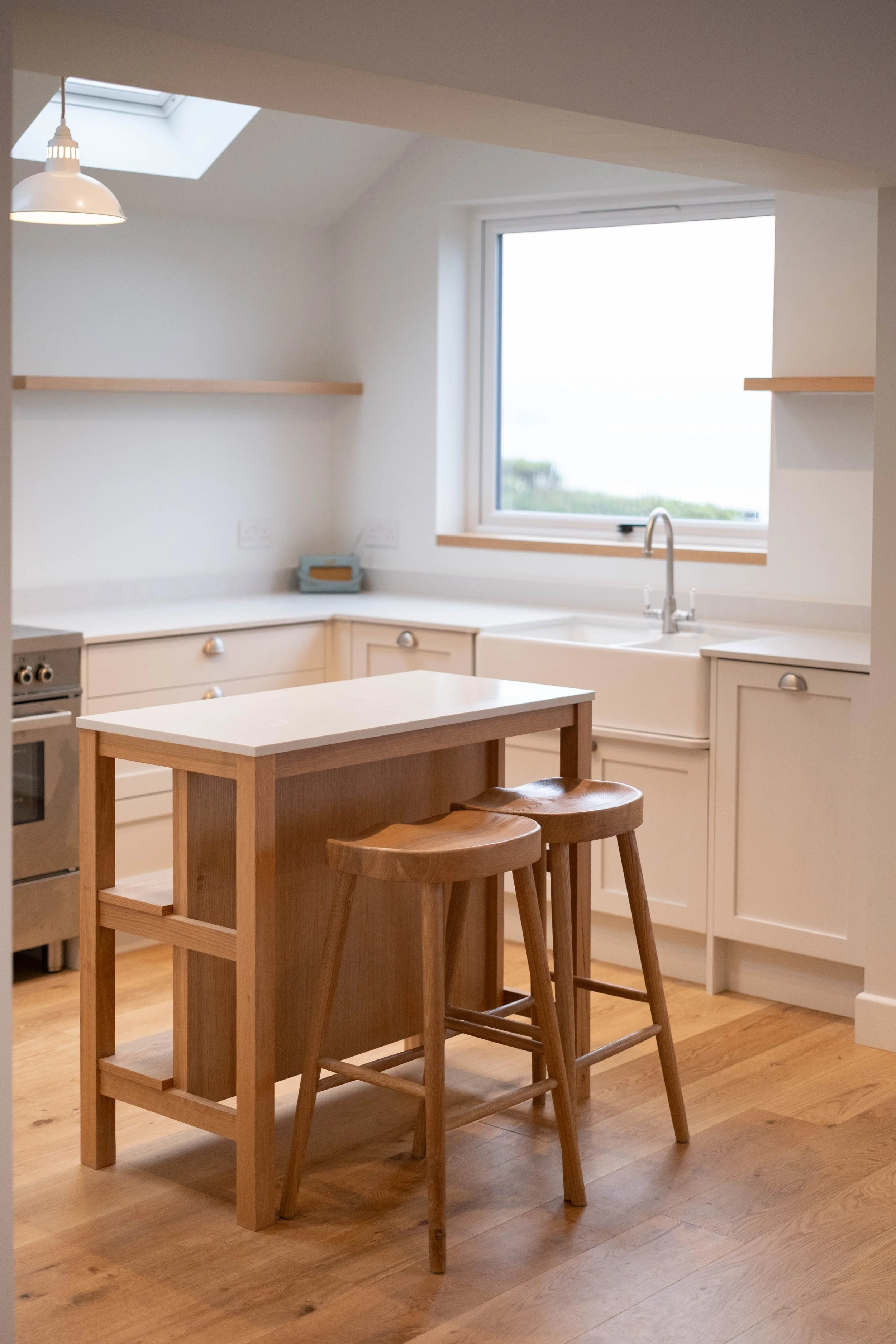 A modern kitchen with white cabinets, a wooden kitchen island with two stools, a large window above the sink, and a skylight in the ceiling. The floor is wooden.