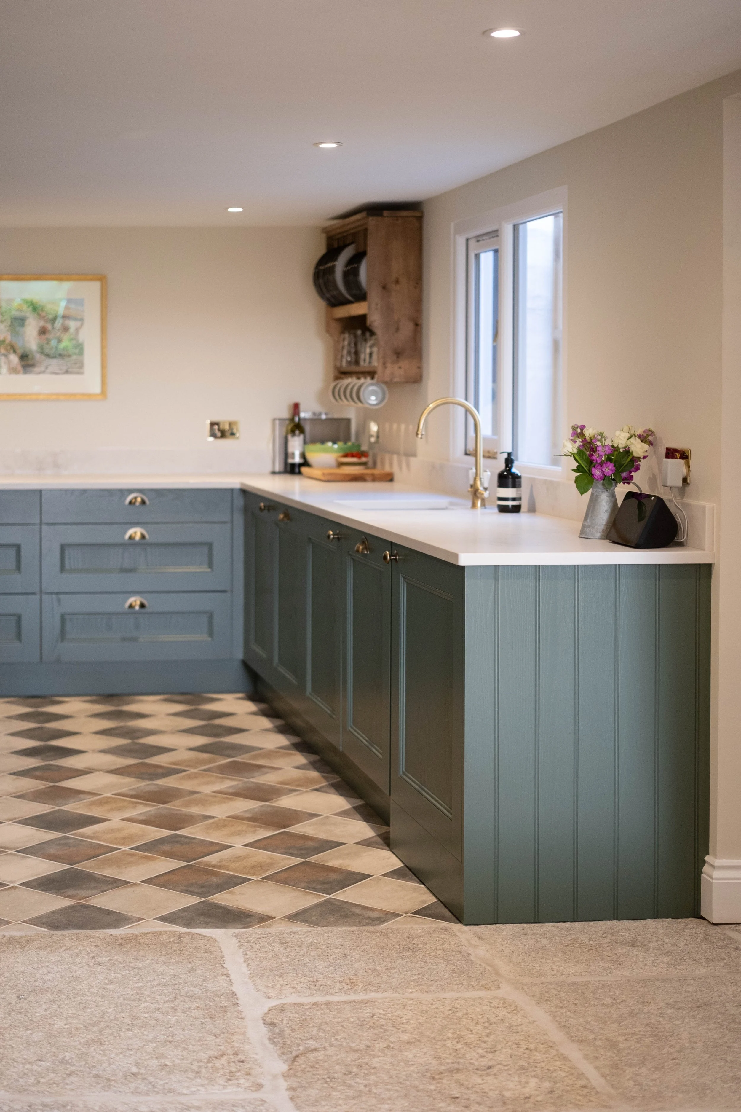 Kitchen with blue cabinets, a white countertop, a gold faucet, a window, and a patterned floor. There are flowers, a black speaker, and a soap dispenser on the counter.