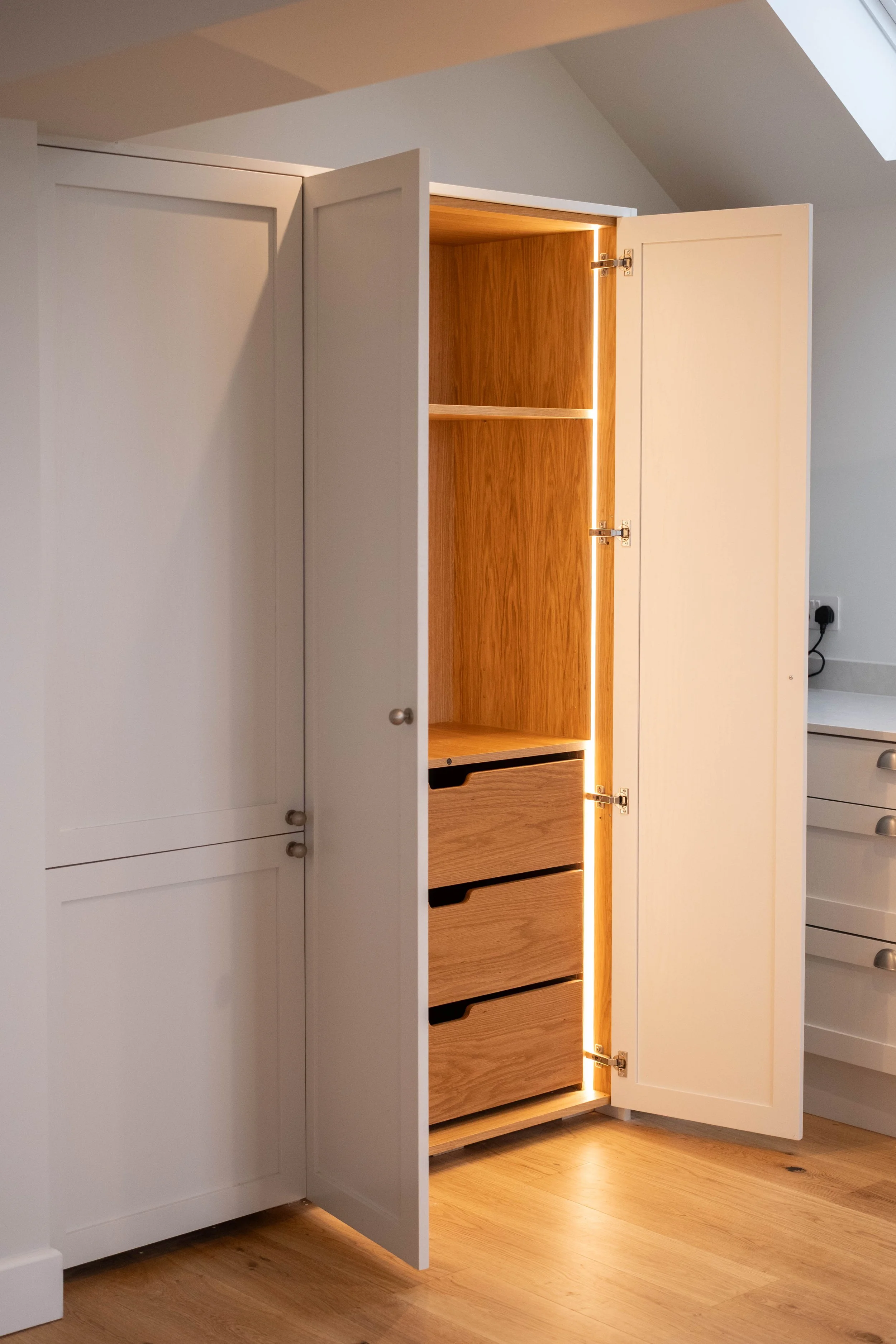 An open white cabinet with wooden interior drawers and shelves, illuminated by a vertical strip of lighting on the right side. There are additional cabinets with silver knobs on the left and right sides of the image.