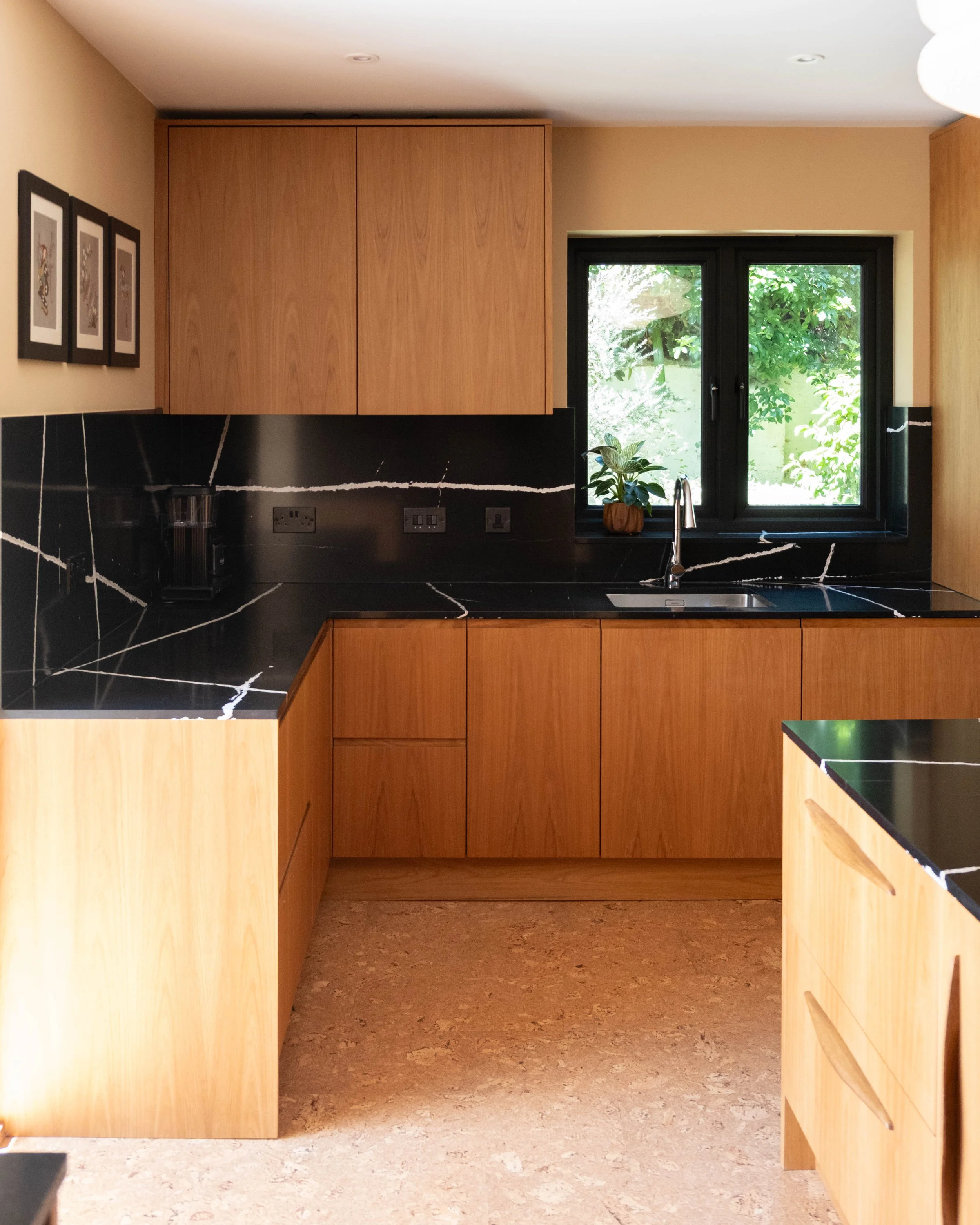 A kitchen with wooden cabinets, black marble countertops with white veins, a stainless steel sink with a faucet, a small potted plant on the windowsill, black-framed double window showing outside greenery, framed art on beige wall, electrical outlets