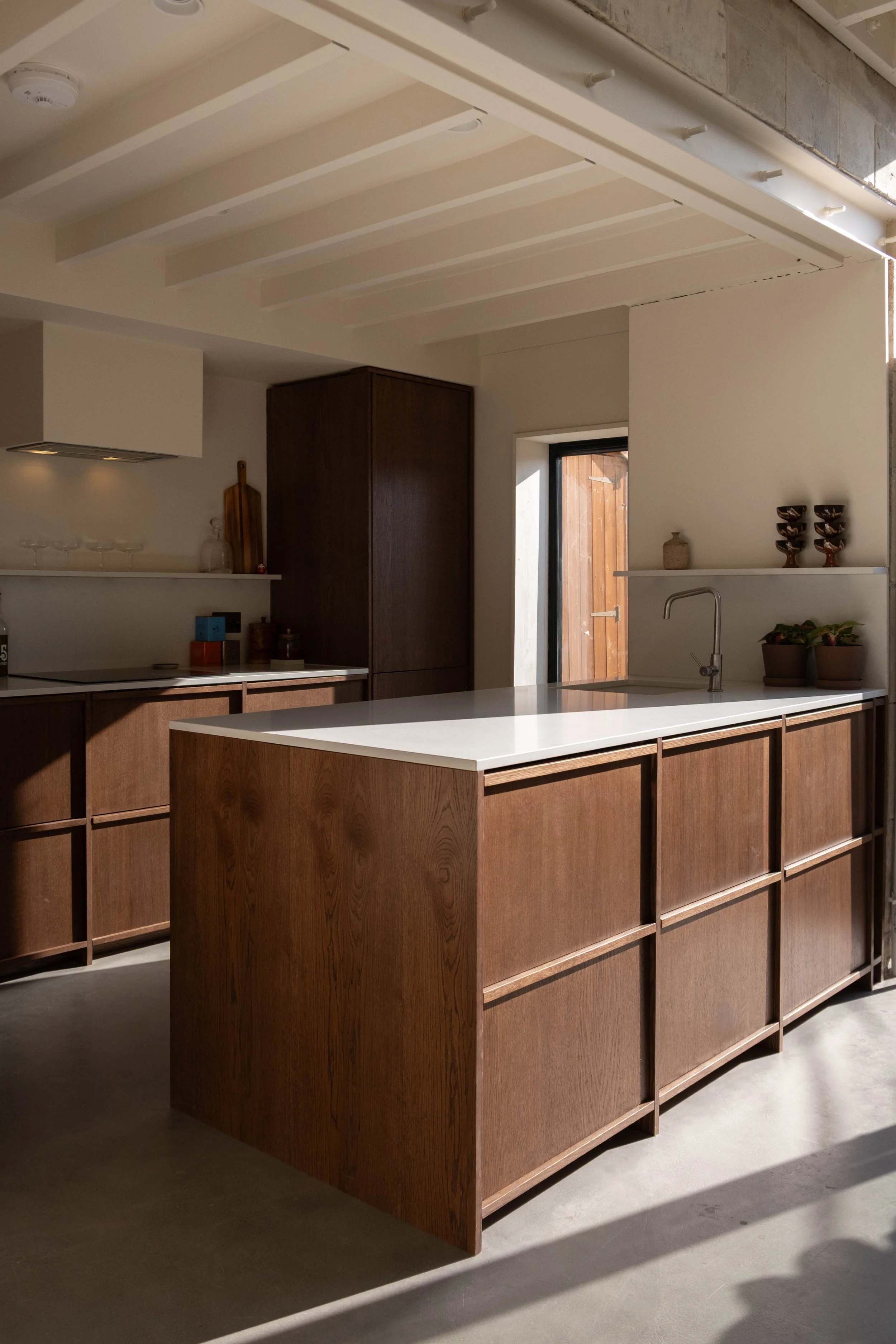 Modern kitchen with wooden cabinetry, white countertops, a black faucet, and decorative bowls and plants on shelves. Light-colored walls and a window with a wooden door outside.