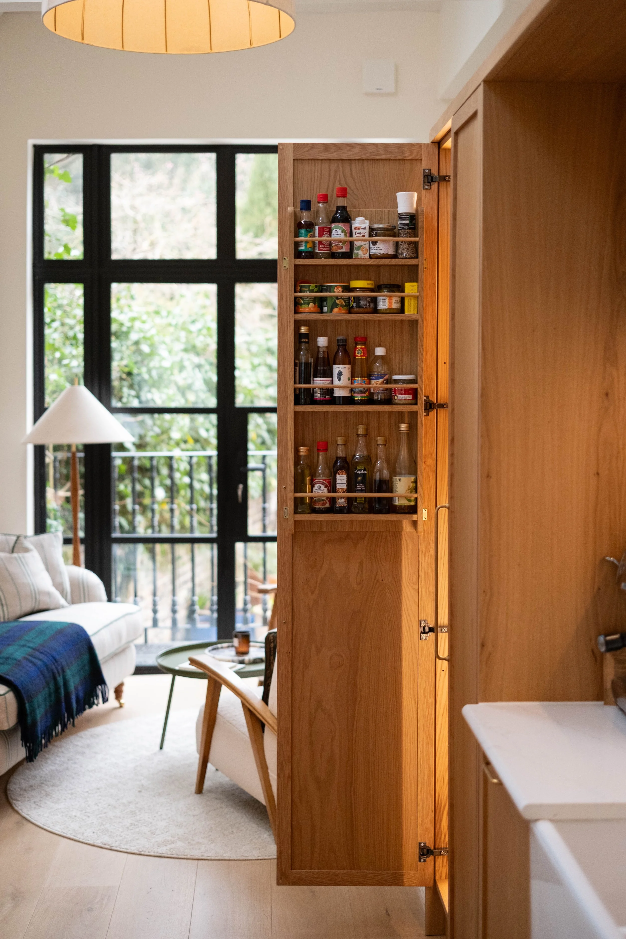 Open wooden kitchen cabinet with spice jars and bottles, view from dining and living area, large window/door with trees outside, living room furniture, and lighting.