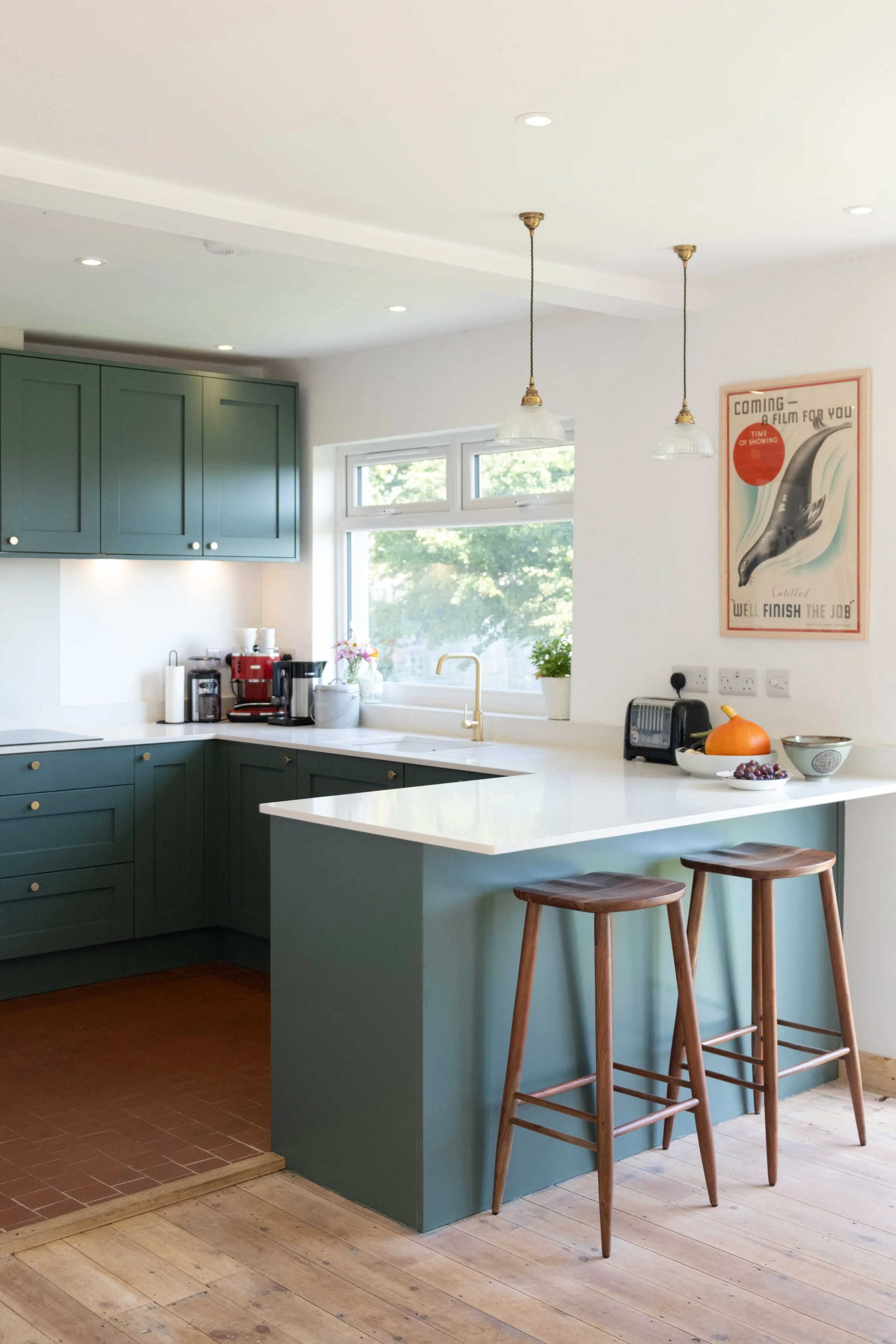 A modern kitchen with green cabinets, white countertops, and two wooden bar stools, with a window showing green trees outside.