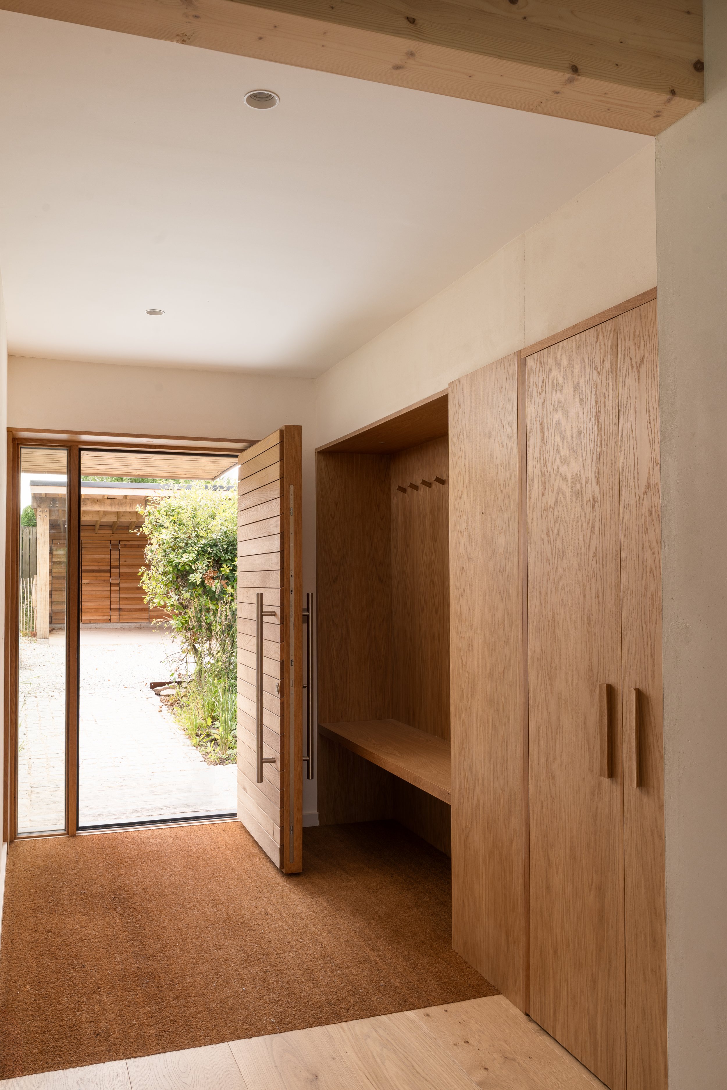 Interior view of a modern home entryway with wooden cabinets, a glass door leading outside, and a garden visible through the door.