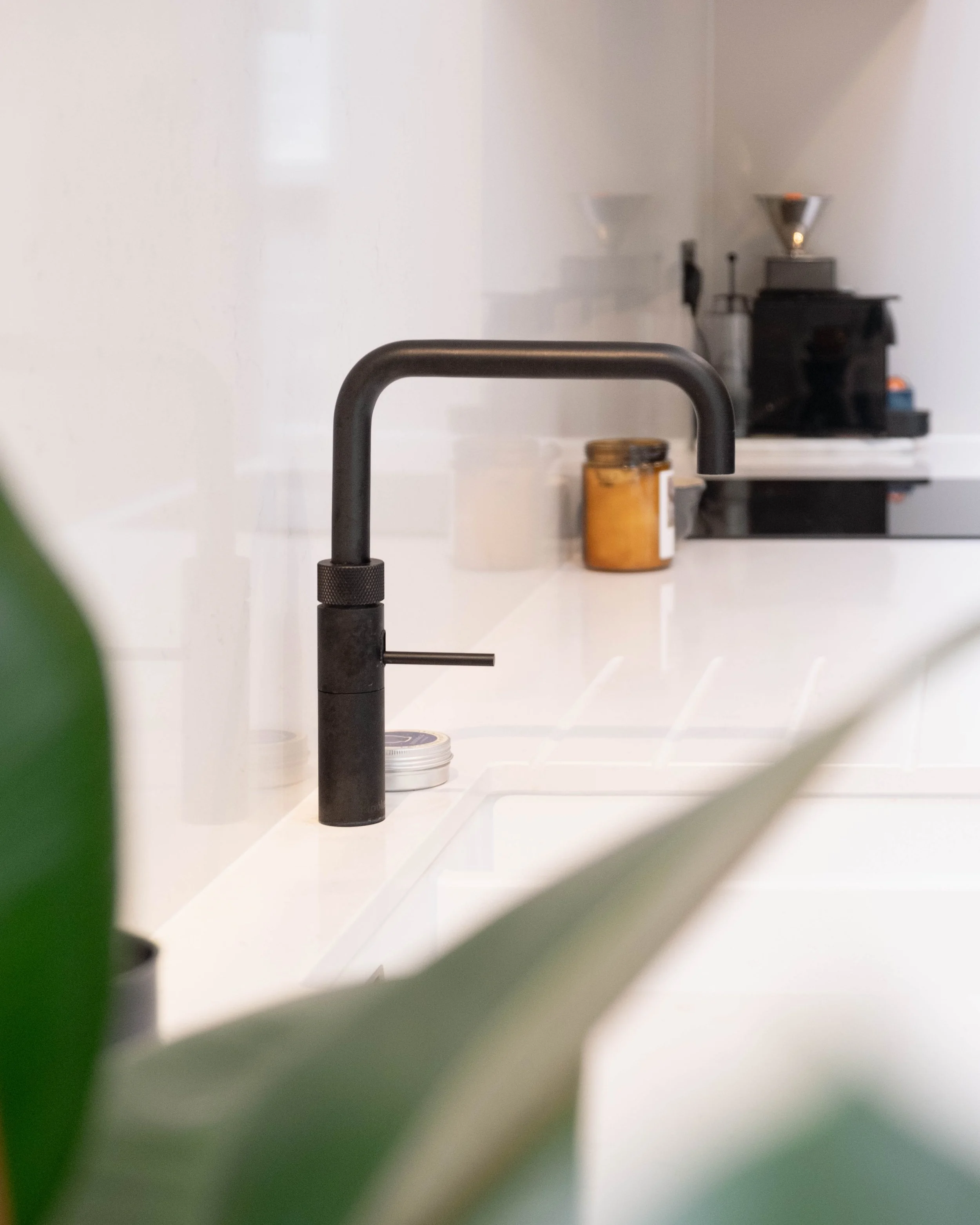 Close-up of a modern black kitchen faucet with a silver handle, on a white countertop. In the background, there is a jar, a coffee grinder, and a coffee filter with orange coffee beans inside, along with a white wall.