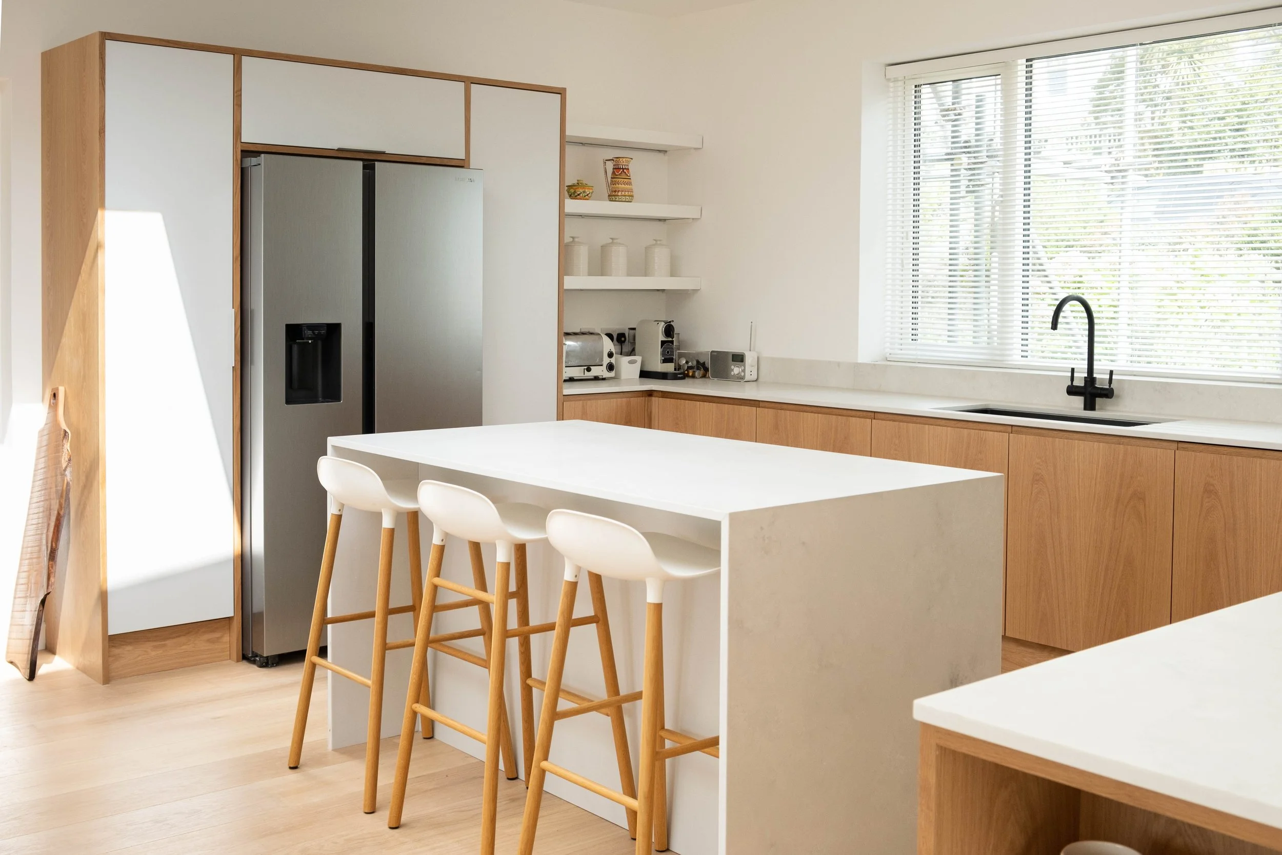 Modern kitchen with white countertops, wooden cabinets, a stainless steel refrigerator, and a large window with blinds.