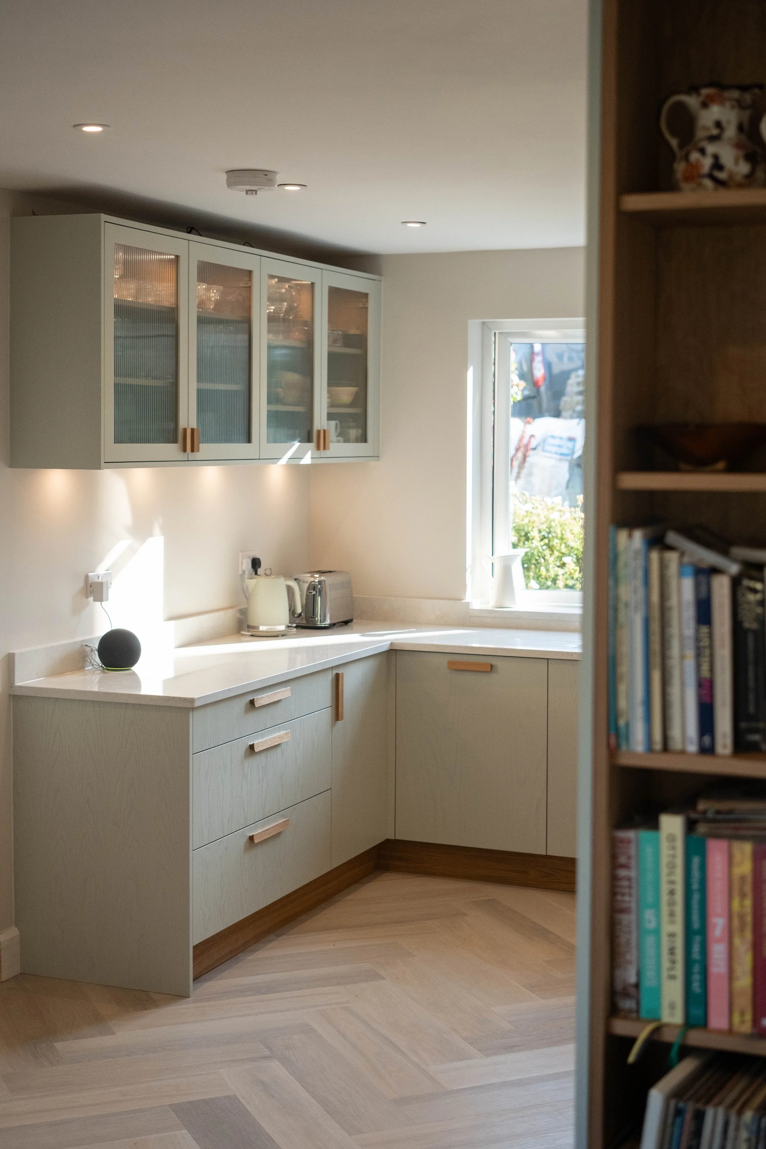 A modern kitchen with light-colored cabinets, a white countertop, and a window letting in sunlight. There are small appliances on the counter, including a kettle and a toaster, with a bookshelf visible on the right side of the image.