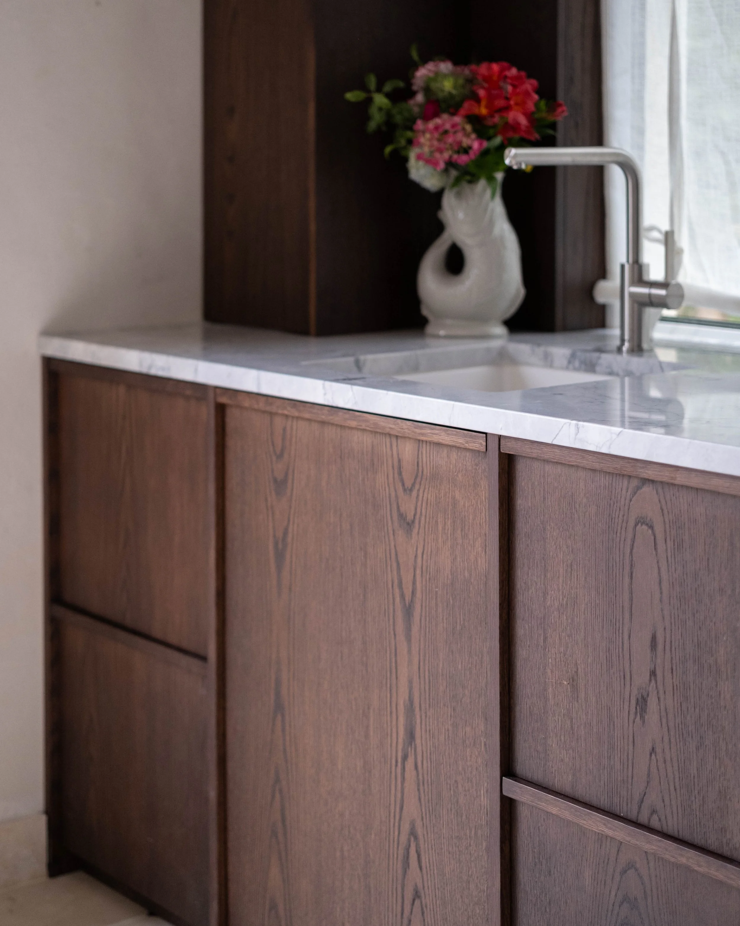 A kitchen sink with a modern silver faucet on a marble countertop. In the background, there is a vase with pink, white, and red flowers placed on a dark wooden surface, next to a window with sheer curtains.
