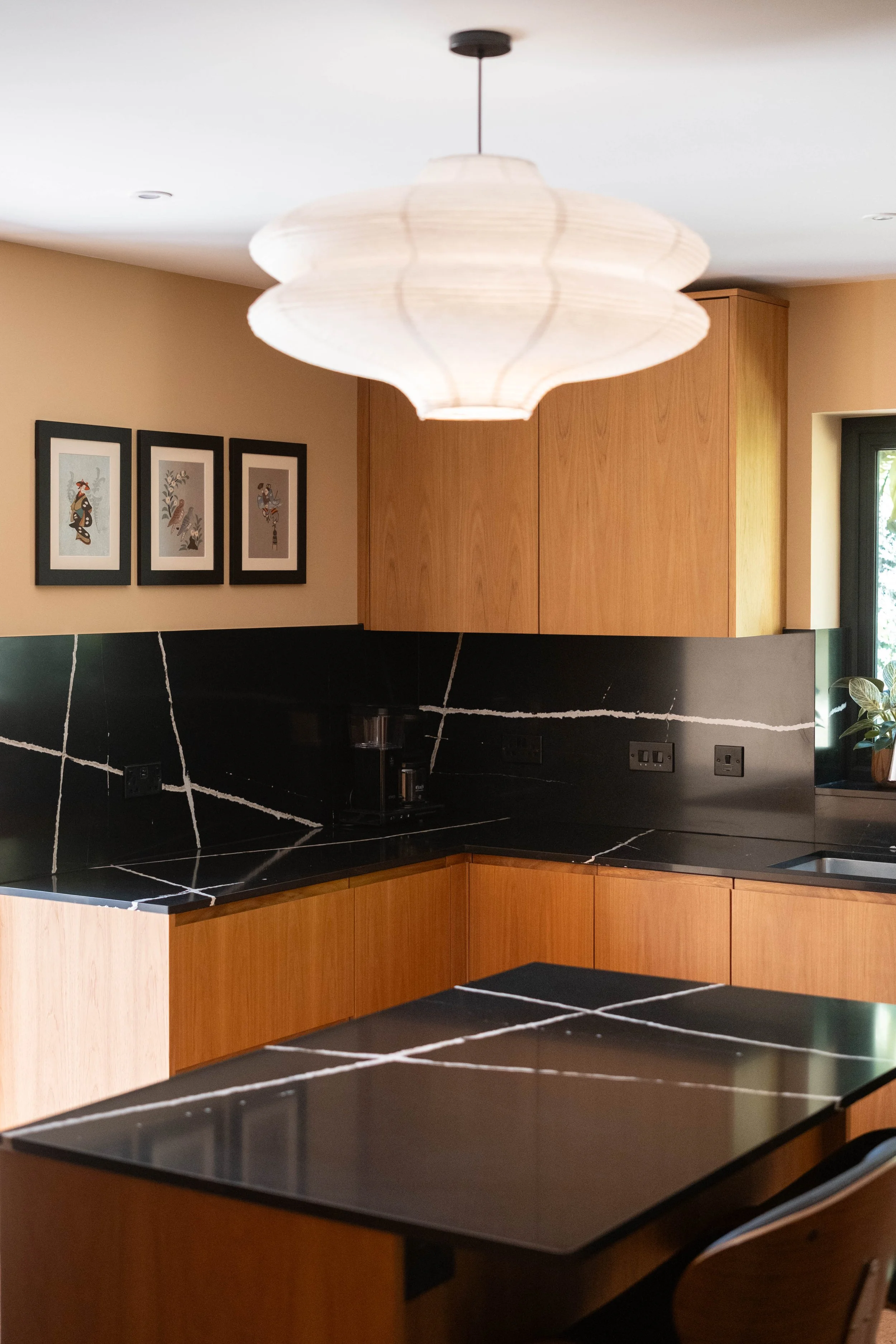 Modern kitchen with black marble countertops, wooden cabinetry, three framed art pieces on beige wall, large pendant light, and window with greenery outside.