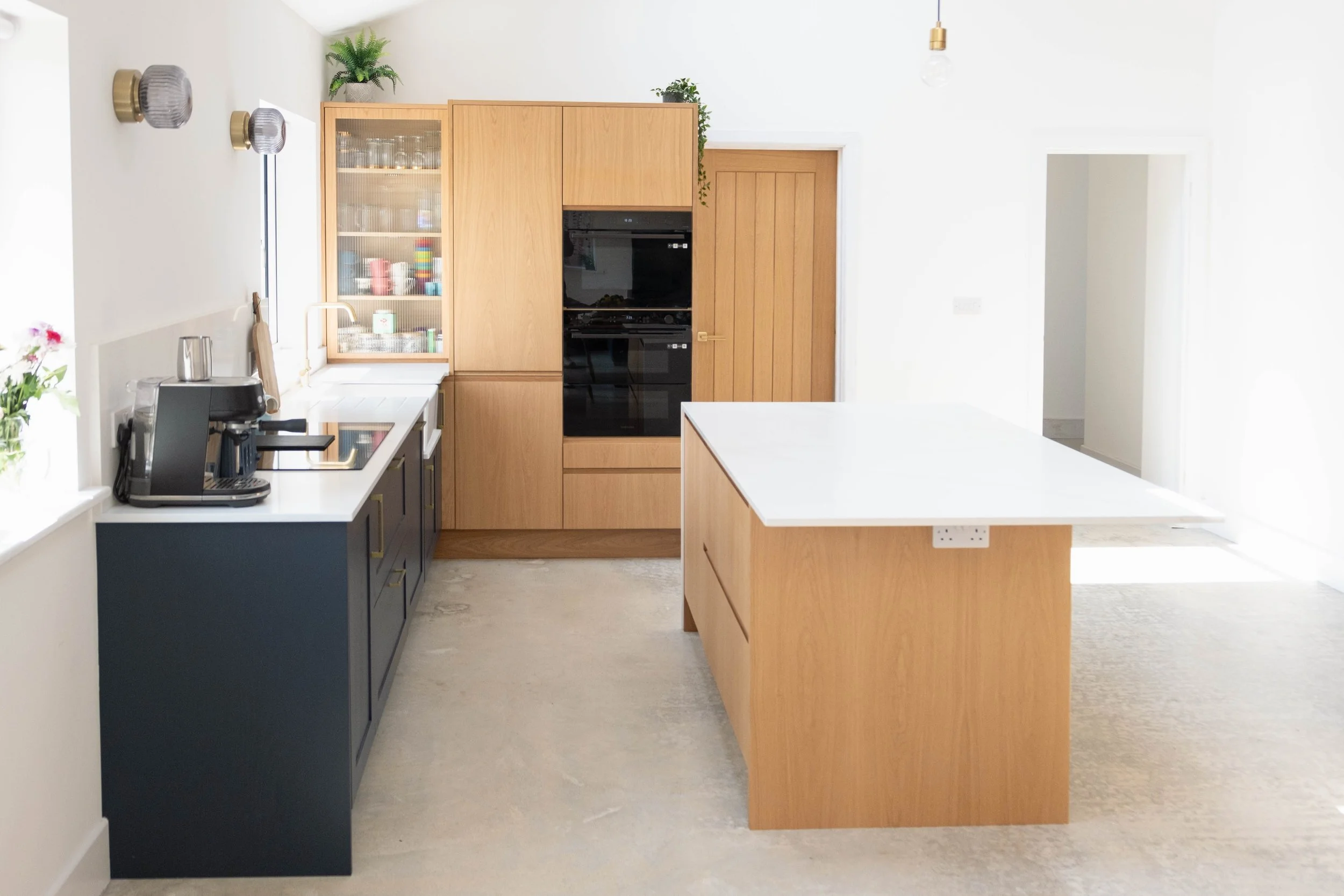 A modern kitchen with a white countertop island, wooden cabinets, black oven, and a black cabinet with a coffee machine on it. There are light fixtures on the wall and plants on top of the cabinets.