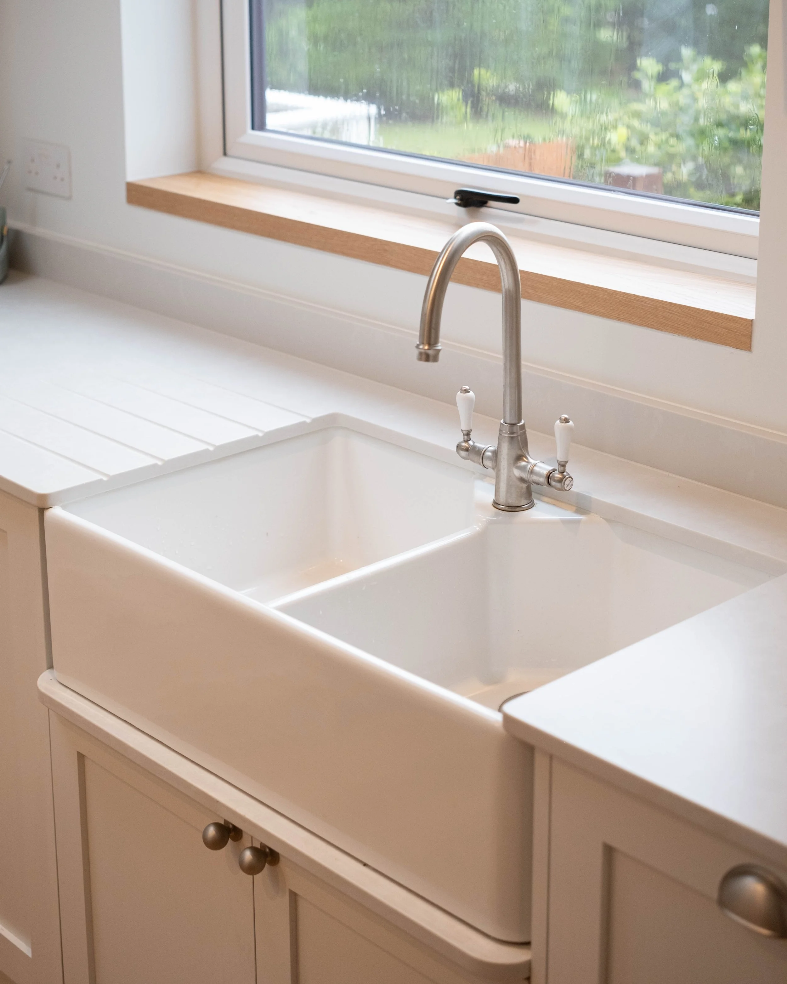 Kitchen sink with a window above, showing a garden outside. The sink is made of white material, and there is a stainless steel faucet. The window has a wooden sill and a handle, with water droplets on the glass. The lower cabinets are white with roun
