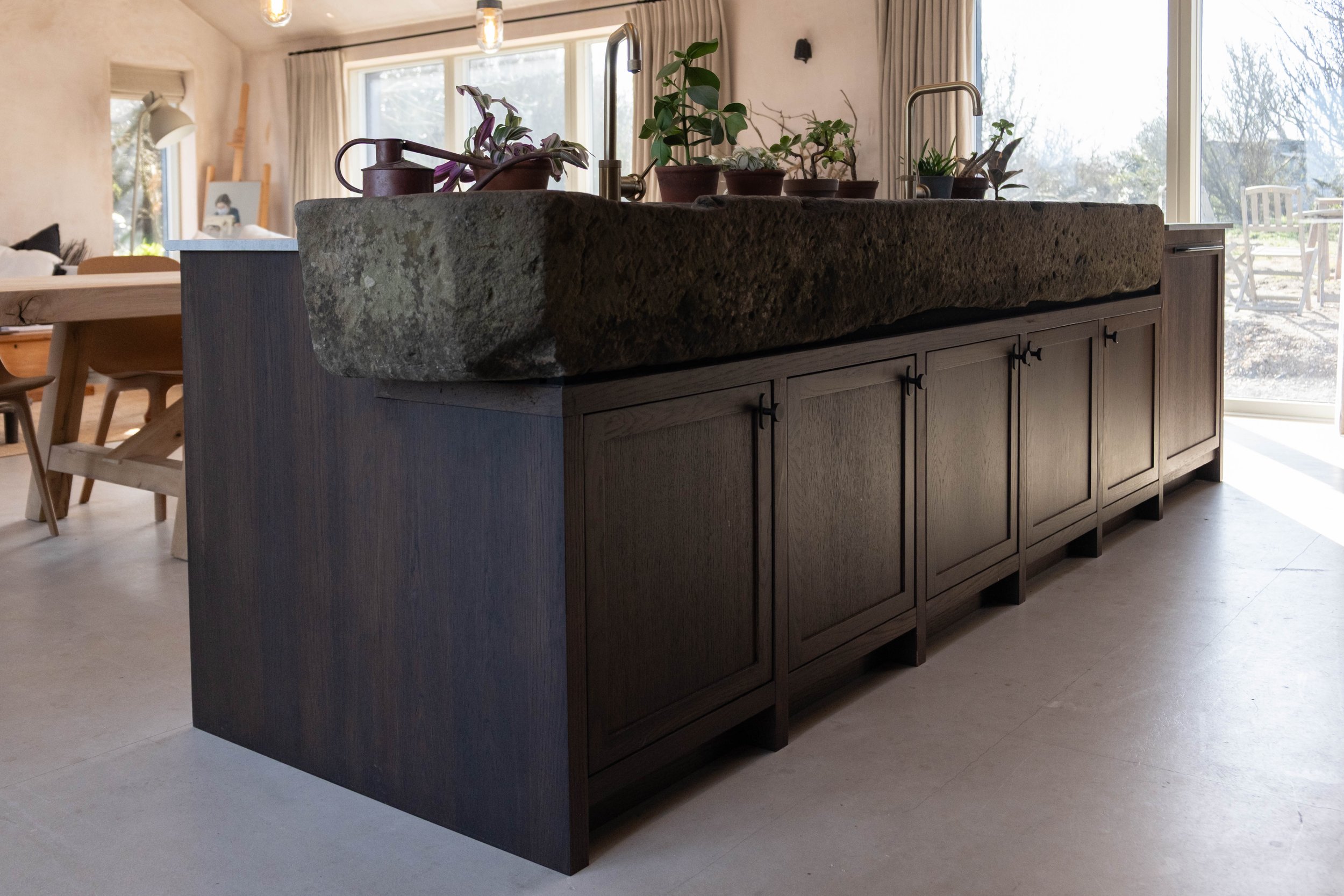Modern kitchen island with a rough stone top and dark wood cabinets, decorated with potted plants. Bright natural light from large windows in the background.