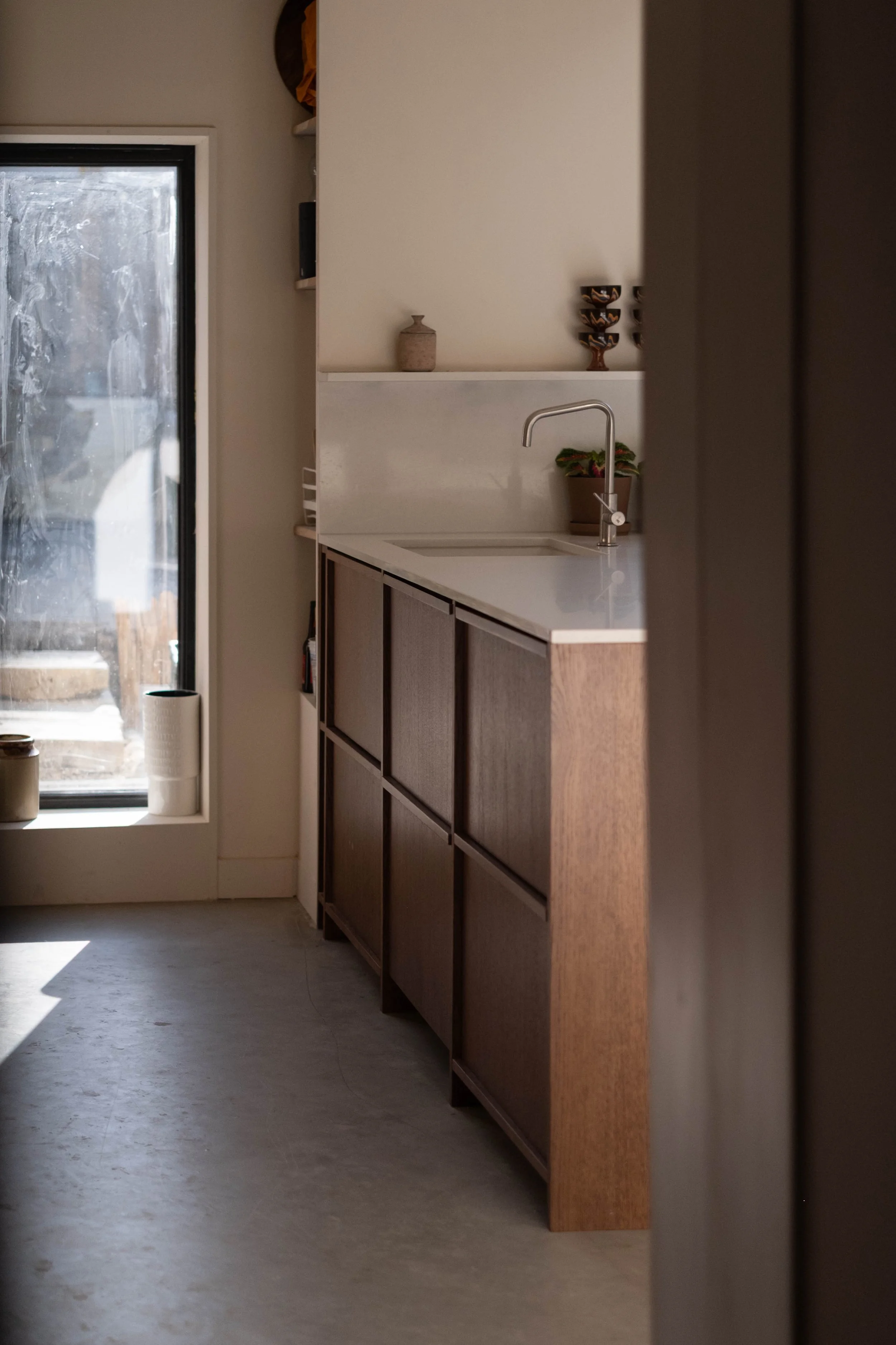 A kitchen with a white countertop, a brown cabinet, a potted plant, a soap dispenser, and decorative bowls on a shelf. Part of a sliding glass door with frosted glass is visible, letting in natural light.