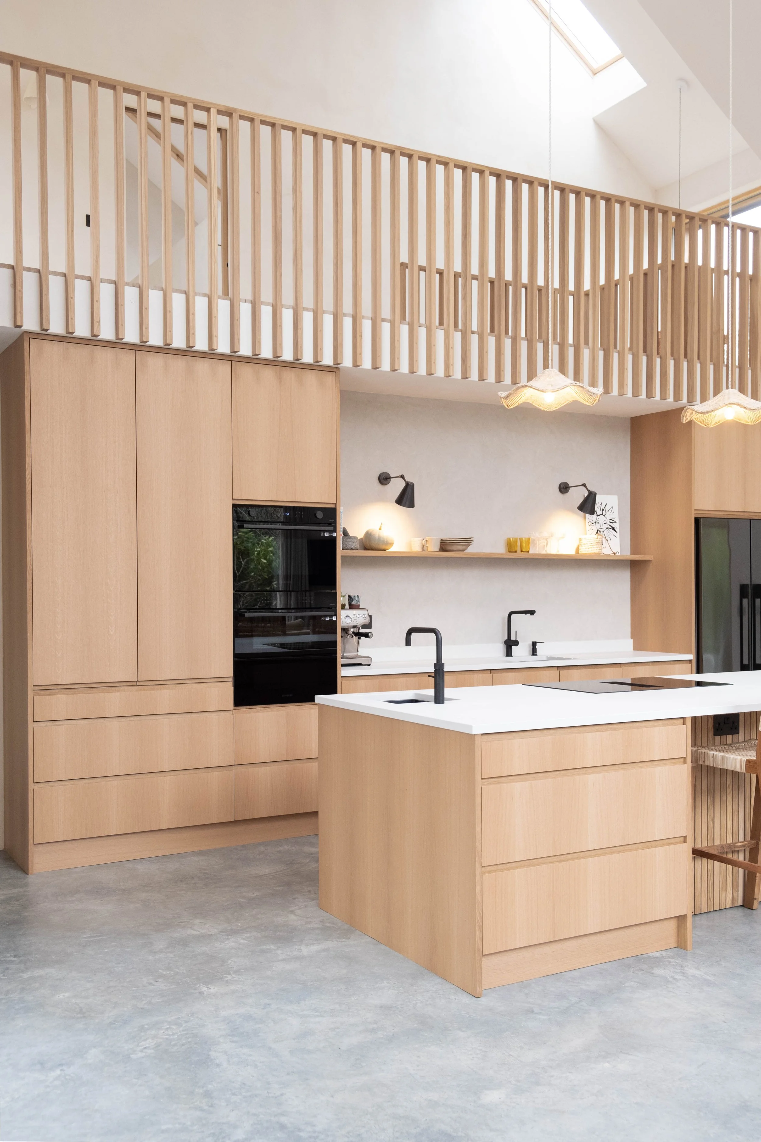 Modern kitchen with light wood cabinetry, a white kitchen island with black faucet, wall-mounted lamps, and minimalistic decor. Loft with wooden railing and skylights in the background.