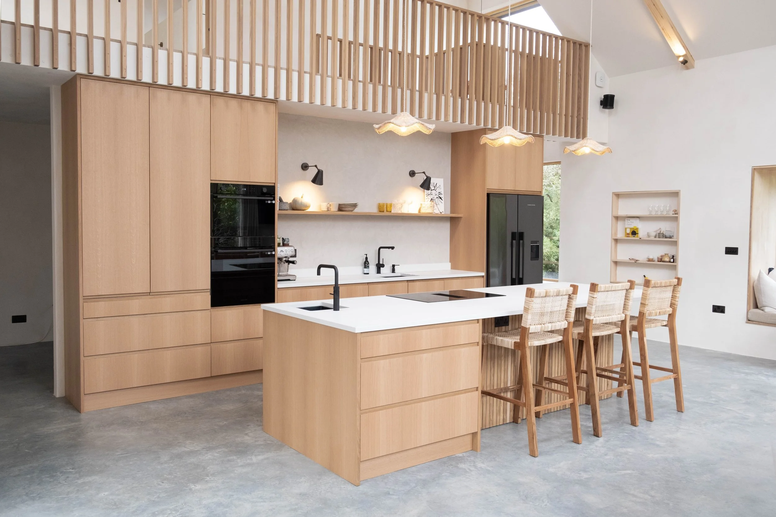 Modern kitchen with light wood cabinetry, white island with black sink and faucet, and wooden chairs at a breakfast bar. Ceiling lights and black wall-mounted lamps provide lighting. Large window and bookshelf visible.
