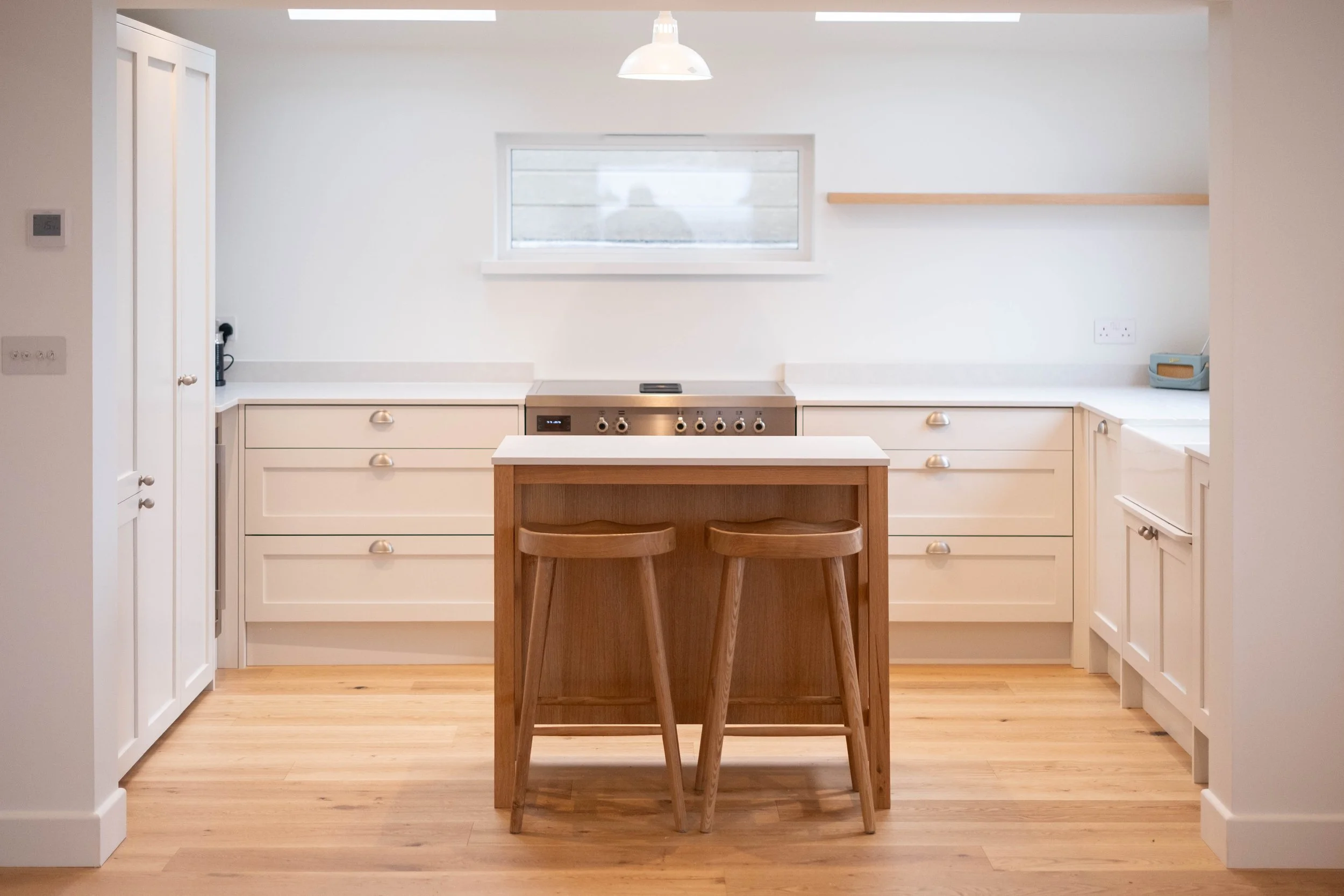 A minimalist kitchen with white cabinets, a central wooden island, and a small window above the stove. There are two wooden barstools positioned in front of the island. The lighting includes a white pendant lamp hanging from the ceiling. The floor is