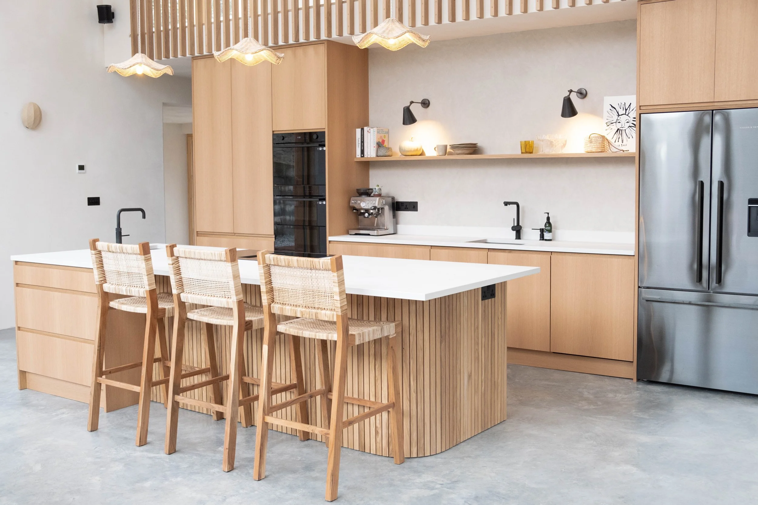 Modern kitchen with light wood cabinets, a large white island, and four wooden chairs with woven seats. Includes a built-in oven, coffee machine, black kitchen faucet, and a fridge. Decorative items on open shelves and wall lighting.