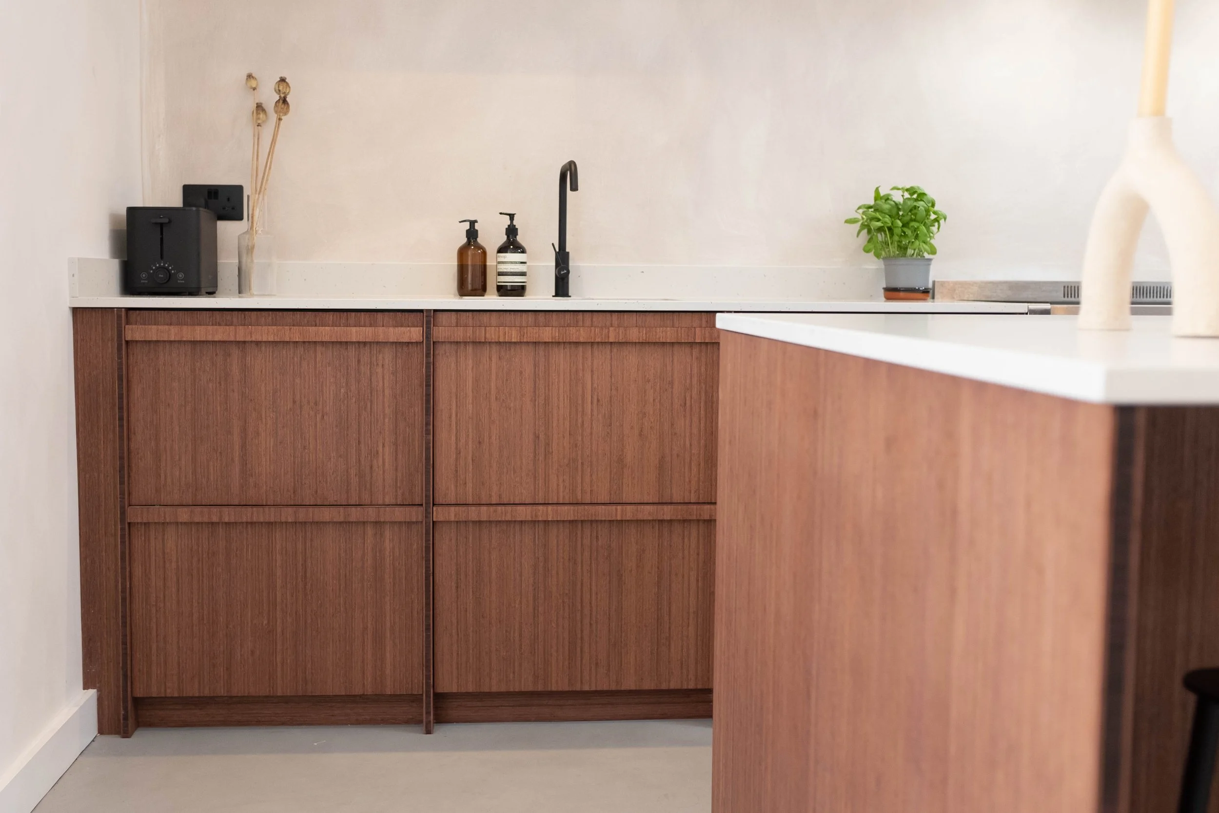 A modern kitchen with wooden cabinets, a white countertop, and decorative items including a potted plant and a ceramic sculpture, with a black faucet and soap dispensers.
