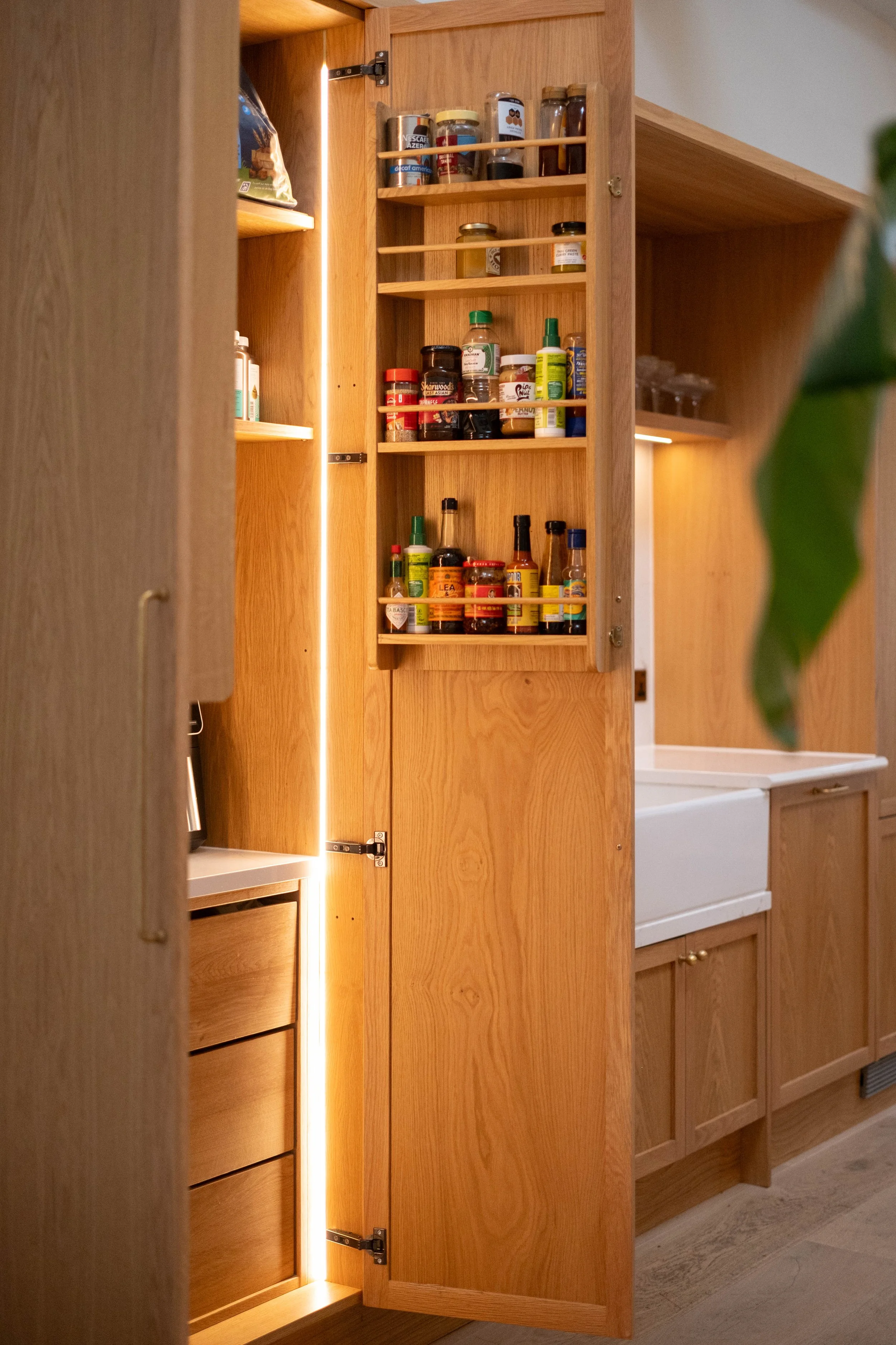 Open wooden kitchen cabinet with shelves holding various jars, bottles, and containers, with a vertical strip of integrated lighting on the left side.