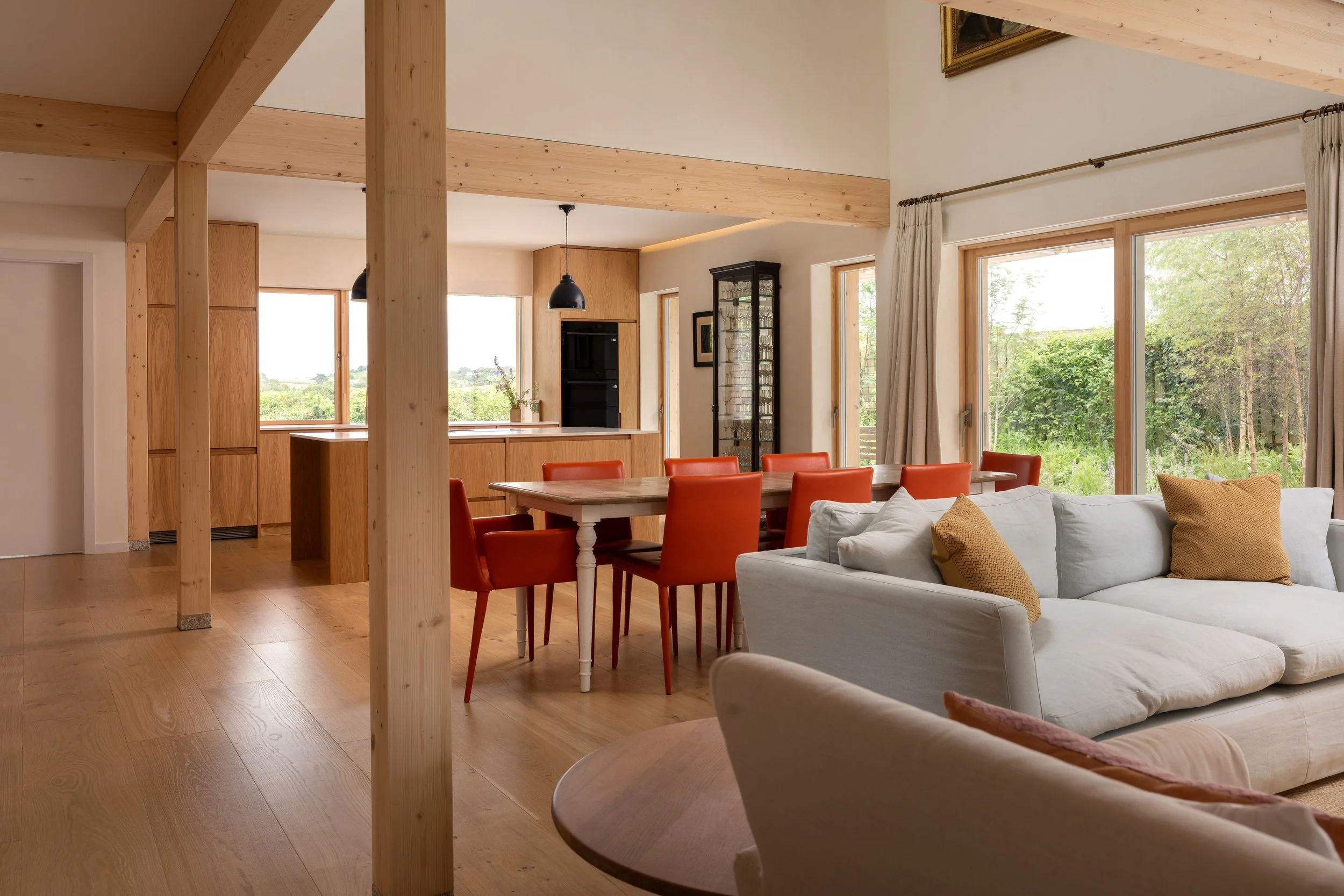 A well-lit open living and dining area with large windows showing greenery outside. The room features wooden accents, including a ceiling beam, paneling, and flooring. There is a white couch with pillows, a wooden dining table with red chairs, and a 