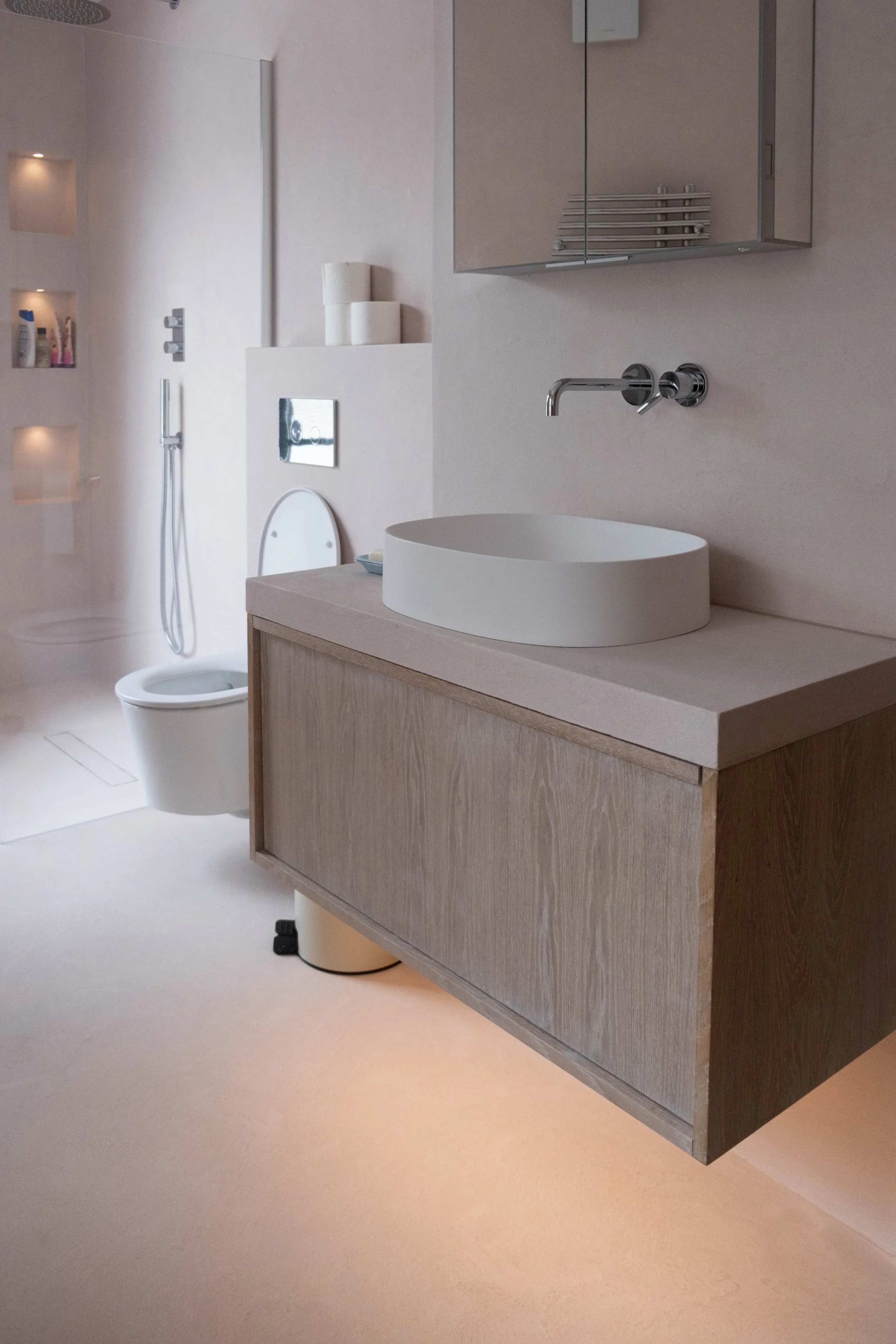 Modern bathroom with a white oval vessel sink on a light wood vanity, a wall-mounted faucet, and a mirror cabinet above. To the left, a toilet with a rectangular flush plate and a shower area with built-in shelves and a handheld shower. The bathroom 