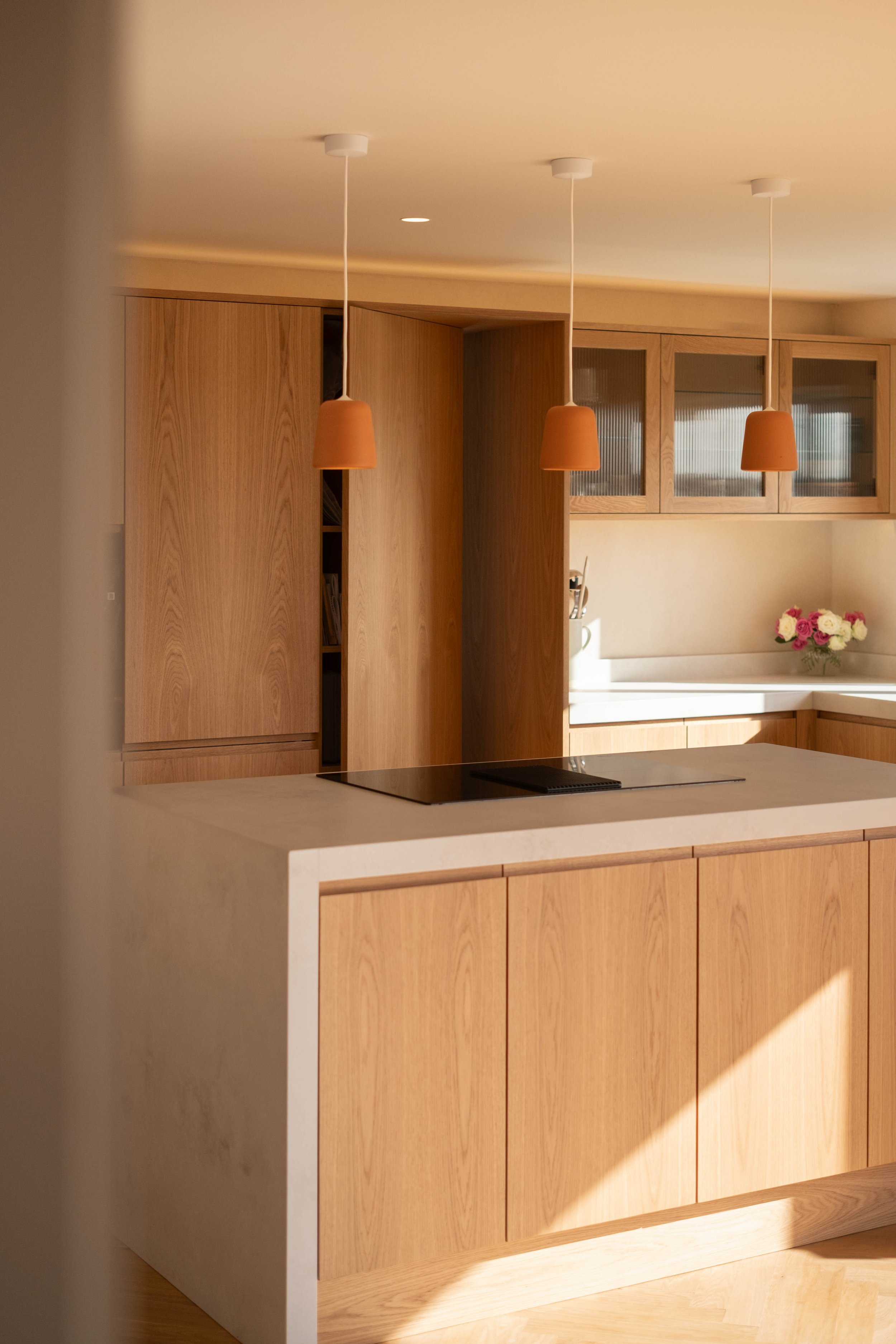 Modern kitchen with wooden cabinets, white countertop, three terracotta pendant lights, and a vase of pink and white roses.