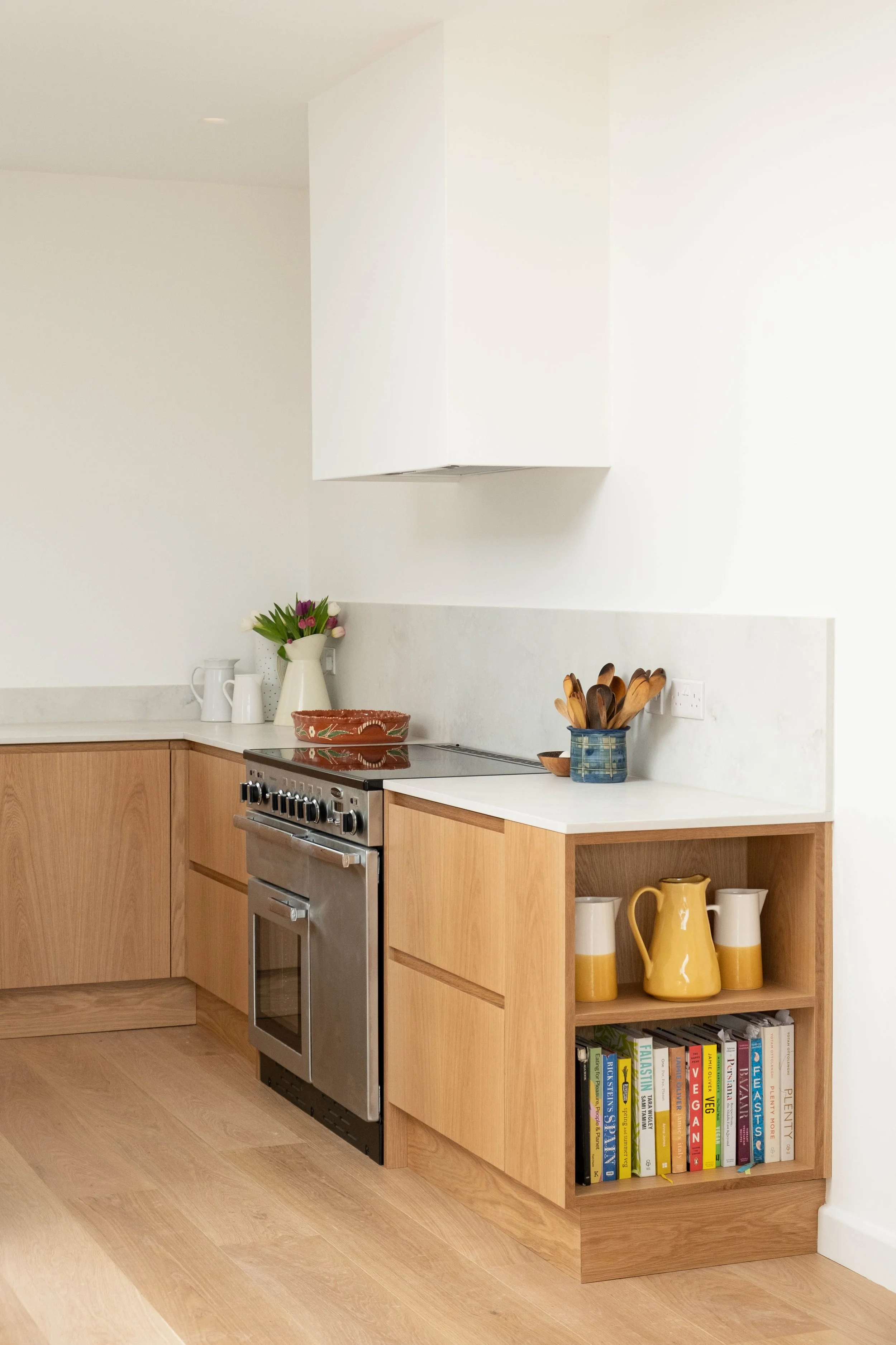 Kitchen corner featuring wooden cabinetry, a stainless steel oven, a white countertop, a small bookshelf with cookbooks, yellow and white ceramics, a potted plant, and wooden utensils in a blue ceramic holder.