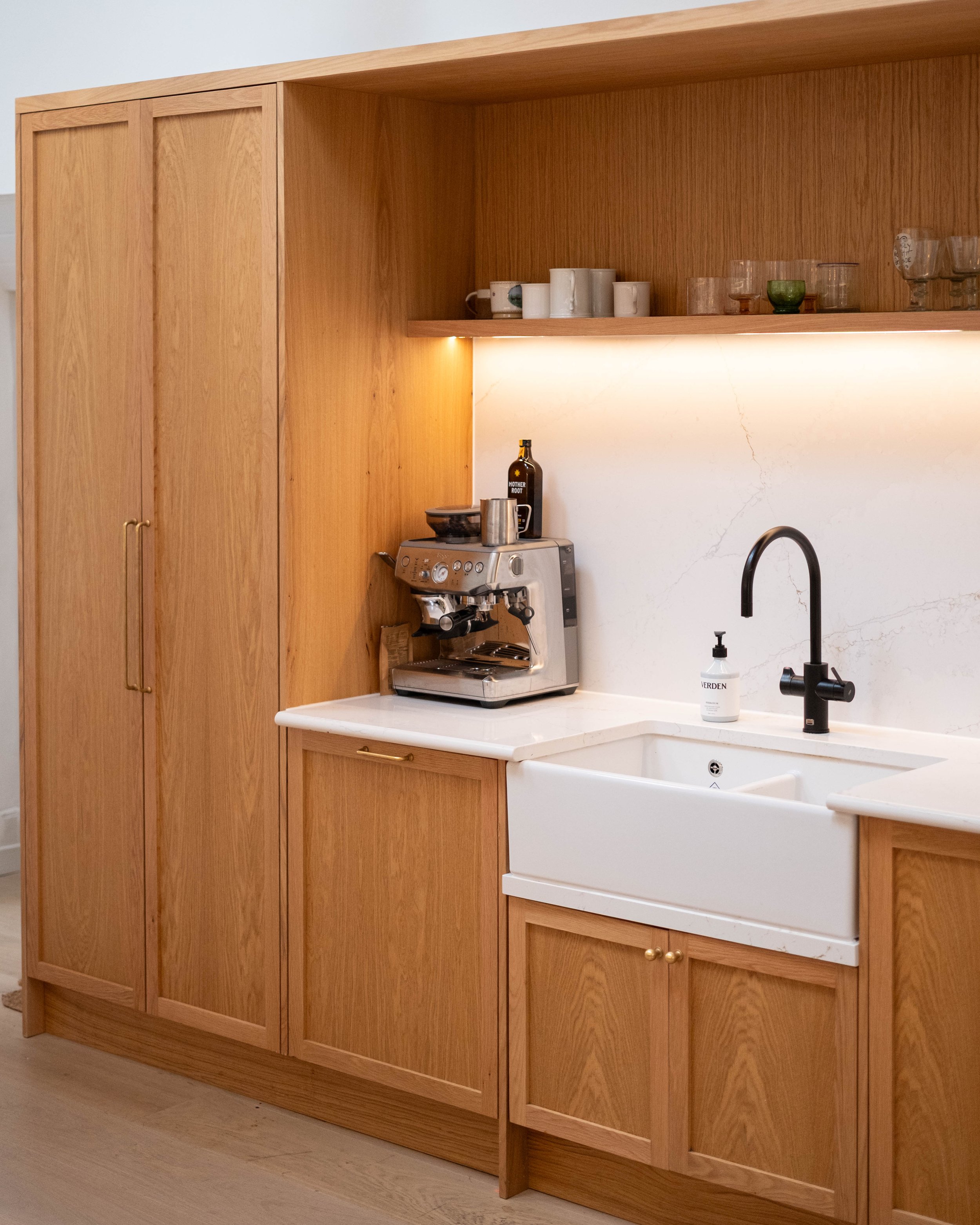 A kitchen countertop with a coffee machine, a bottle labeled 'Mother Root,' a silver mug, a soap dispenser, a black faucet, white marble backsplash, and built-in wooden cabinets with gold handles.