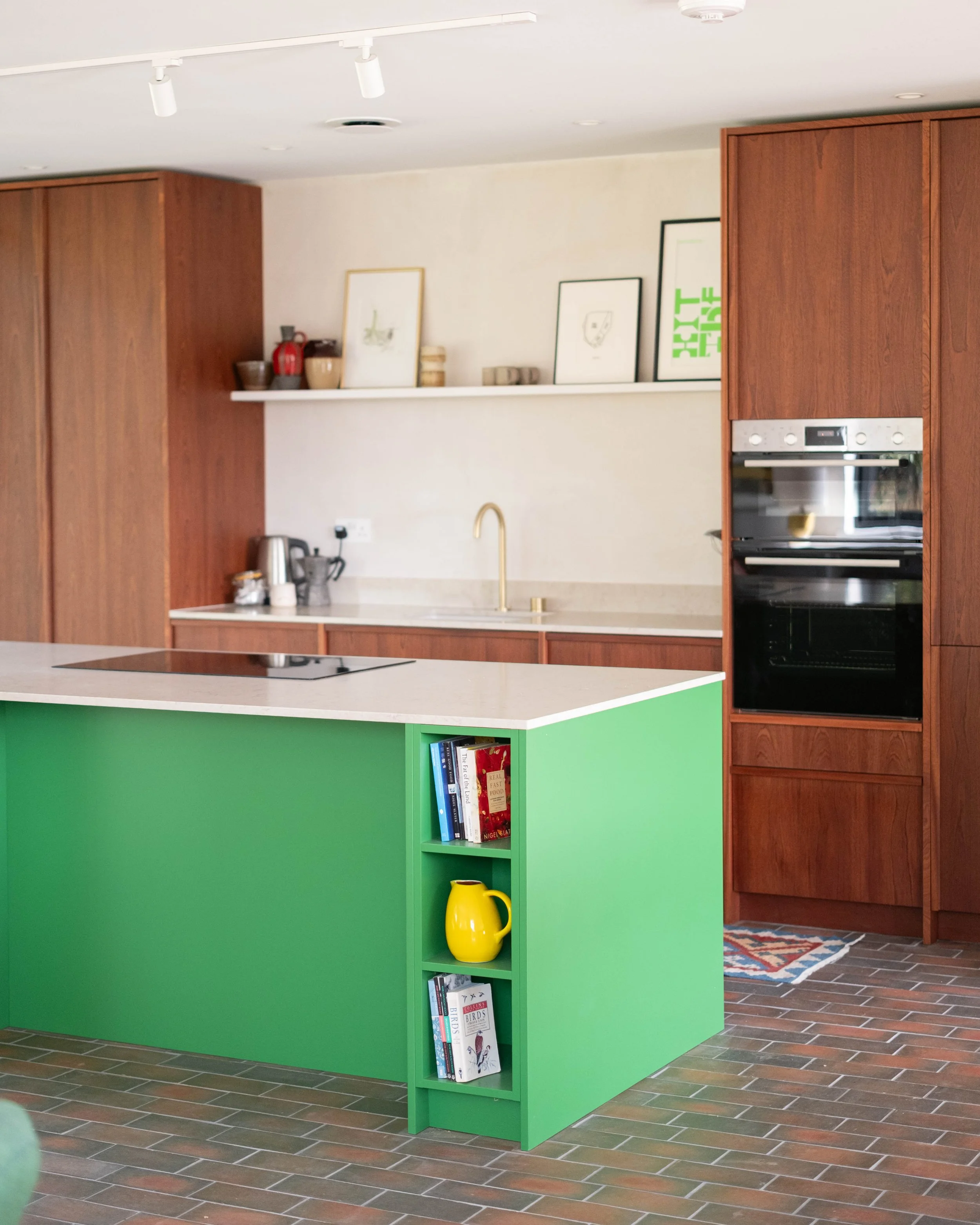 A kitchen with wooden cabinets, a white countertop, and a green bookshelf with books and a yellow jug. There is a built-in oven and a yellow pitcher on the floor. The floor is made of brownish tiles, and there are framed pictures and decorative items