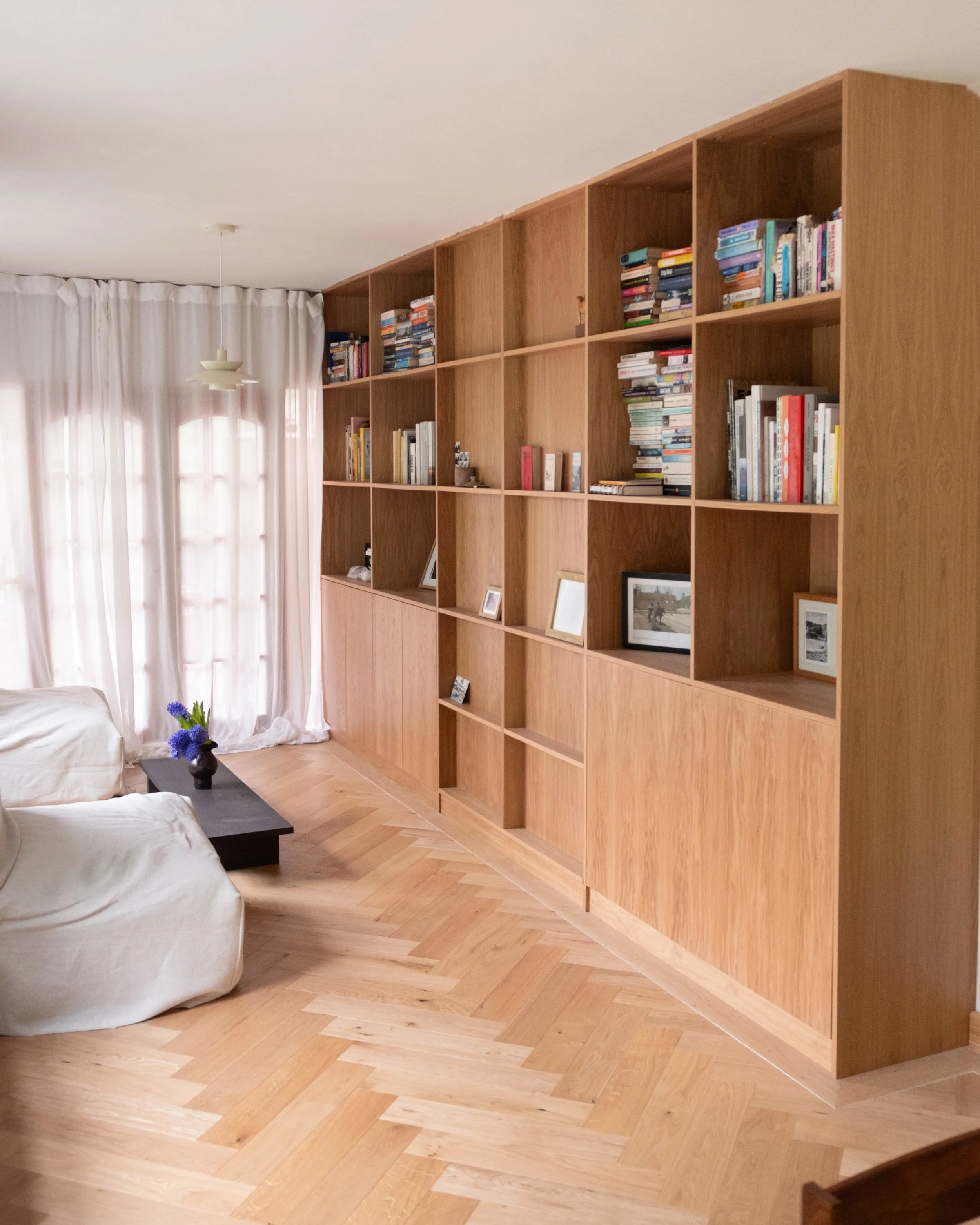 Living room with a wooden bookshelf, white curtains, a couch with white covers, a black coffee table with a small vase of purple flowers, and light-colored wood flooring.