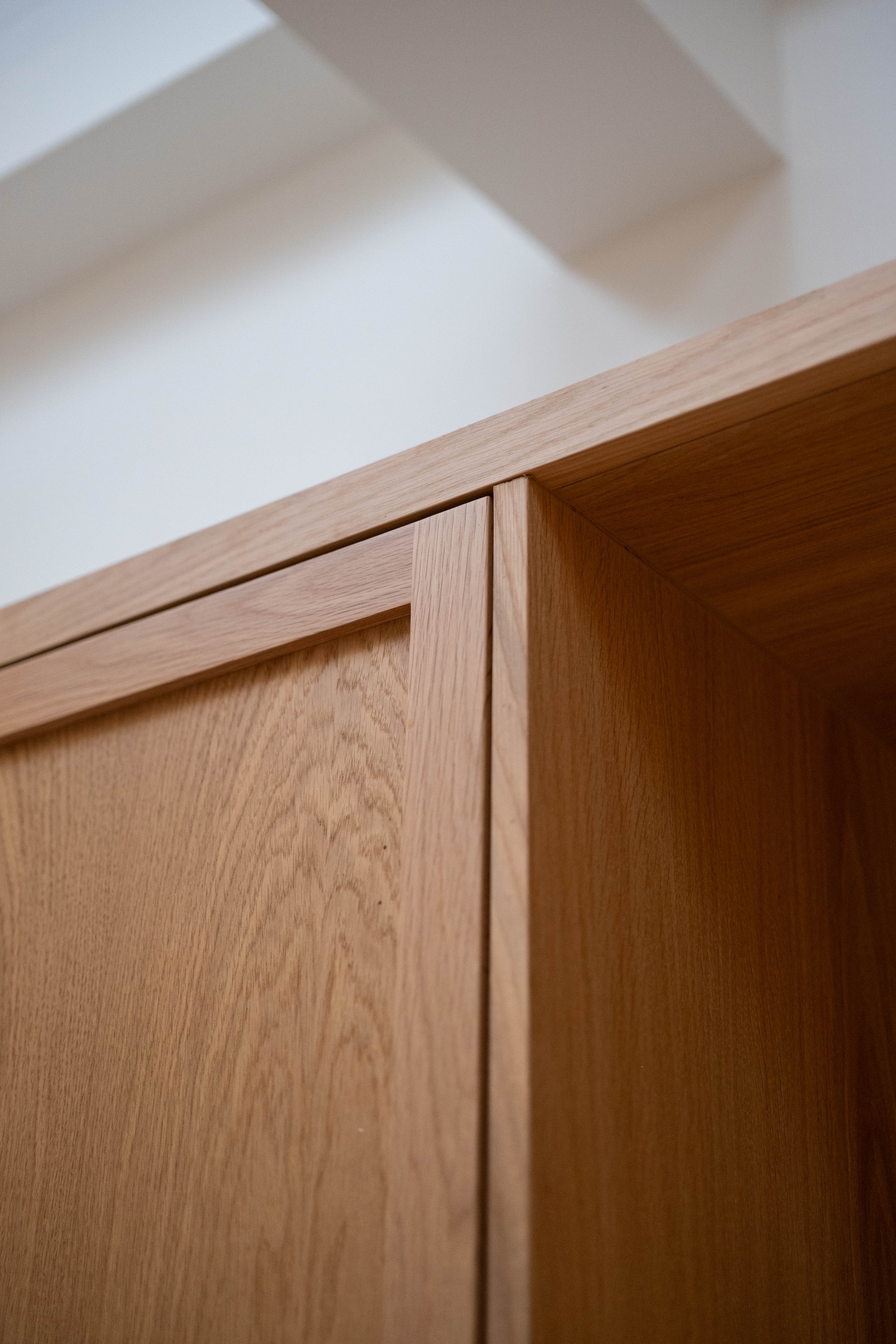 Close-up photo of a wooden cabinet with smooth surface and visible wood grain, featuring a corner and the top edge, against a plain white wall background.