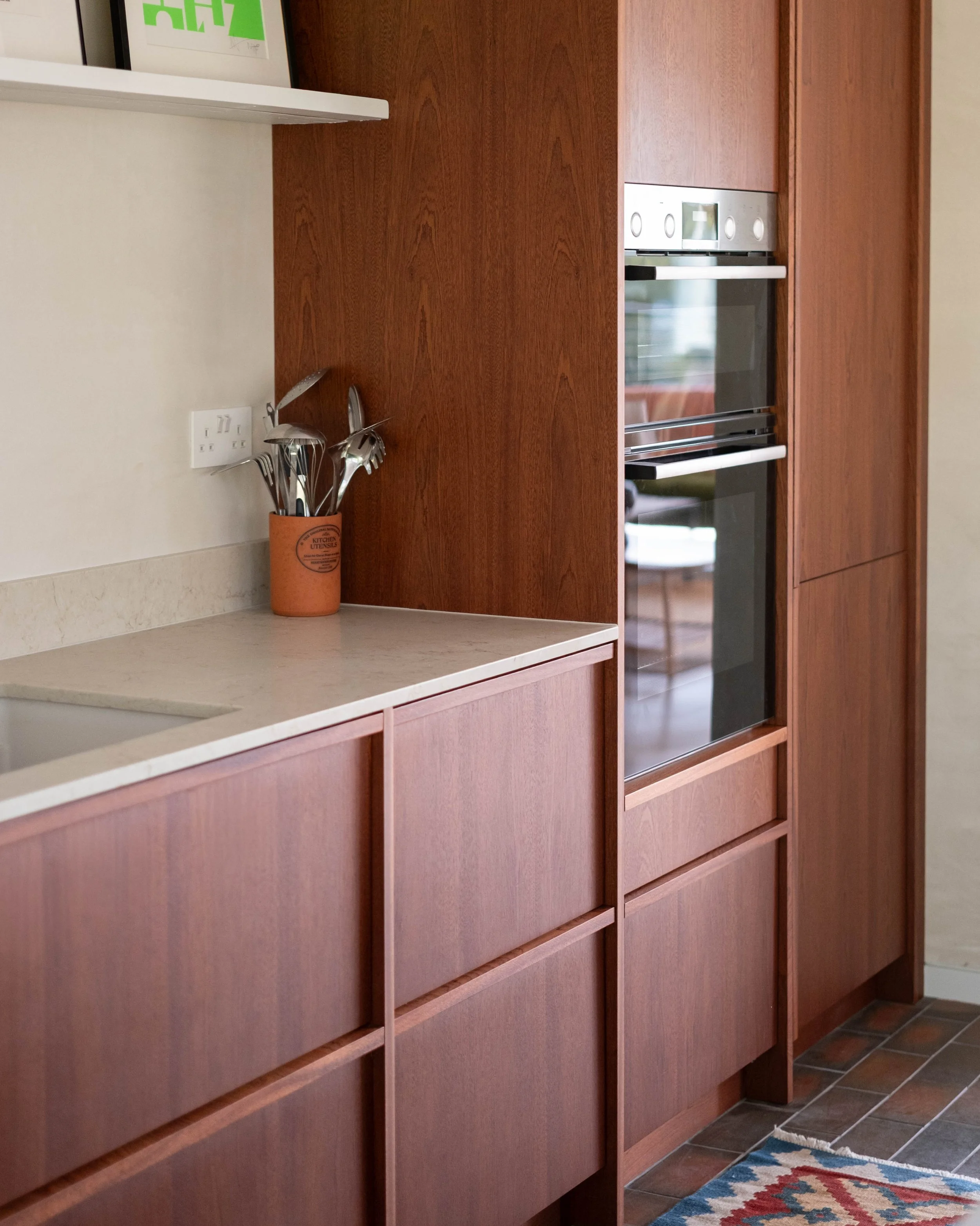 Kitchen with dark wood cabinets, a built-in oven, a small countertop area with a utensil holder, a colorful rug on tile floor, and a framed artwork on the wall.