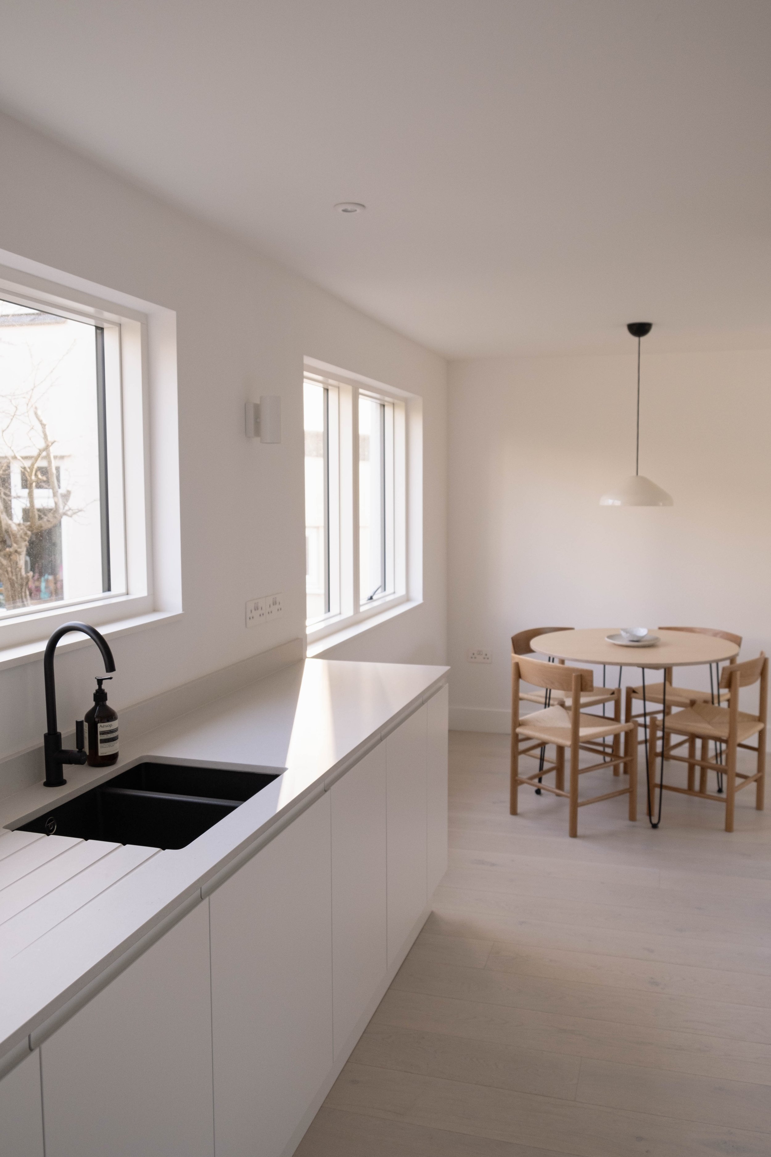 Modern kitchen with white cabinets, black sink, soap dispenser, and large windows, adjacent to a round dining table with wooden chairs and a hanging pendant light.